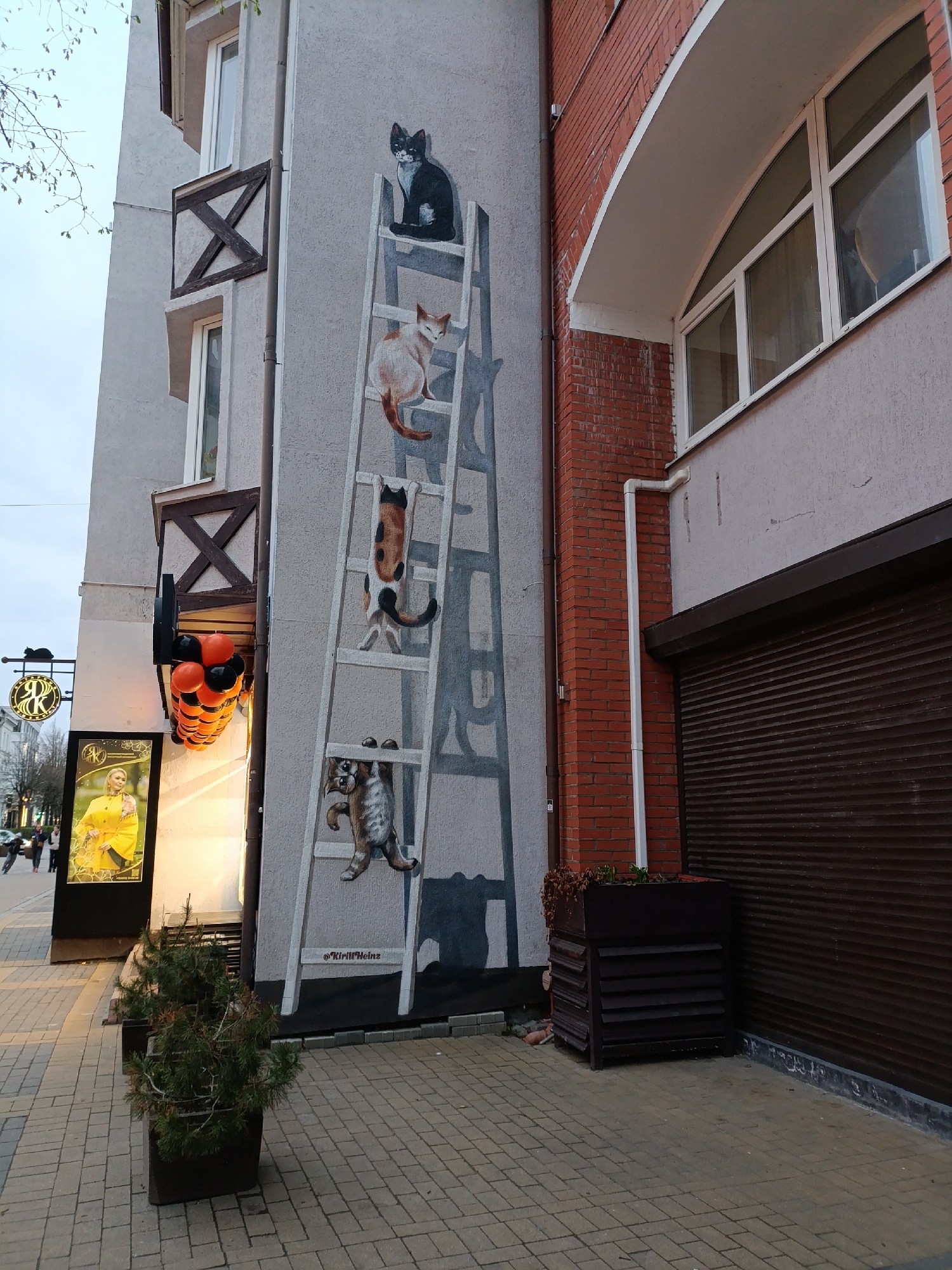 The image captures a building in Zelenogradsk, Russia, notable for a large mural depicting cats climbing a white ladder painted on its exterior wall. The ladder is positioned against a gray wall with the cats strategically placed on different rungs.
To the left, a street scene includes a sign featuring a portrait of a woman in a yellow jacket, and black and orange balloons are affixed to the building’s facade. A small potted evergreen sits at the base of the mural.
Adjacent to the mural, a building with a brick facade features a large arched window above a brown roll-up door. A planter box sits alongside this section of the building. The sidewalk is paved with light-colored bricks. People are visible in the distance.