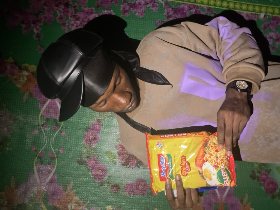 A person wearing a black cap lies on a floral-patterned surface. The individual's eyes are closed. The person wears a brown jacket with a purple design. A watch is visible on their left wrist. The person holds a yellow package of Indomie Chicken Flavor instant noodles. The scene is illuminated with strong shadows.