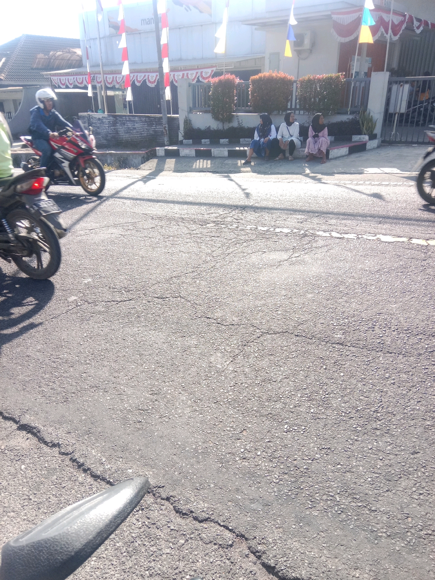 In Kertek, Indonesia, a street scene unfolds with a cracked asphalt road in the foreground. A motorcycle rider in a white helmet and blue jacket speeds past on a red motorcycle. Further down the road, several young women are seated on a raised curb in front of a building adorned with red and white banners and flags, suggesting a festive occasion. The building appears to be a commercial establishment or office. The environment is sunlit, casting shadows on the road.