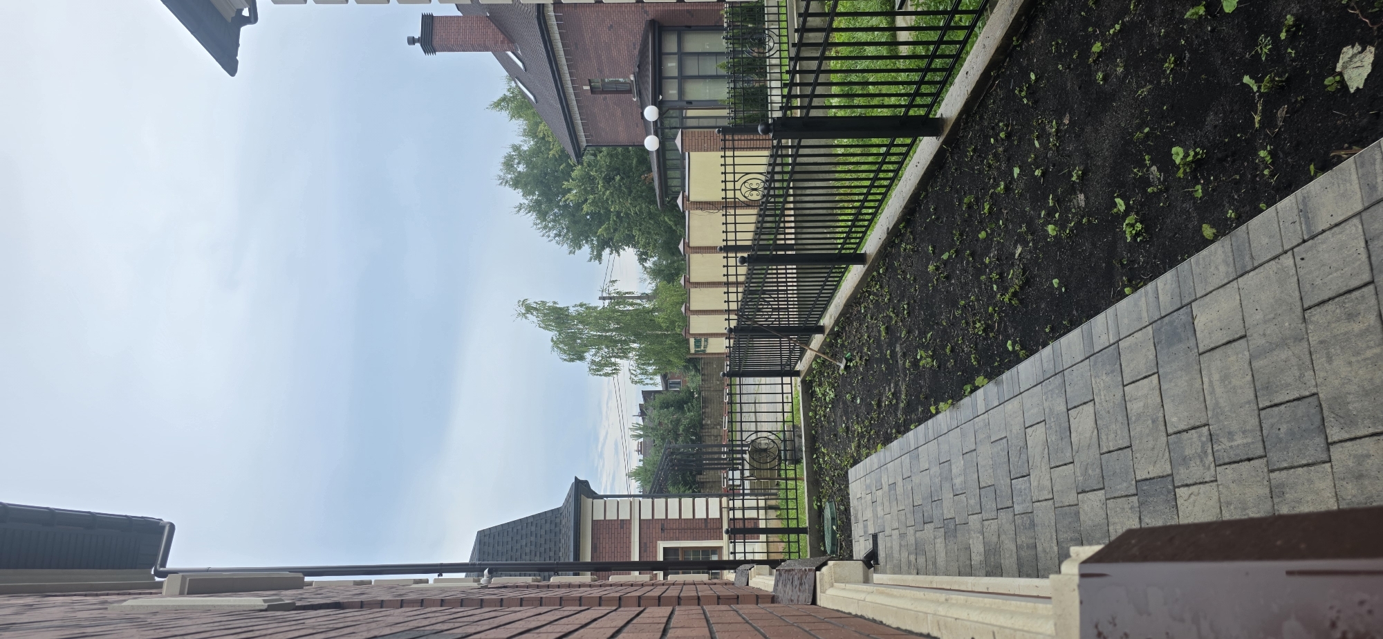 The image shows a pathway leading away from a brick building, bordered by a black metal fence. Beyond the fence is a bed of dark soil with scattered small green plants, suggesting a garden area that is either newly planted or under cultivation. A rake is visible lying in the garden bed. In the background, there are more brick buildings, a light-colored fence, and lush green trees under a cloudy sky. The overall scene depicts a residential outdoor area, likely in a suburban or rural setting. The location is identified as Русская Деревня-2 Кп, Russia.