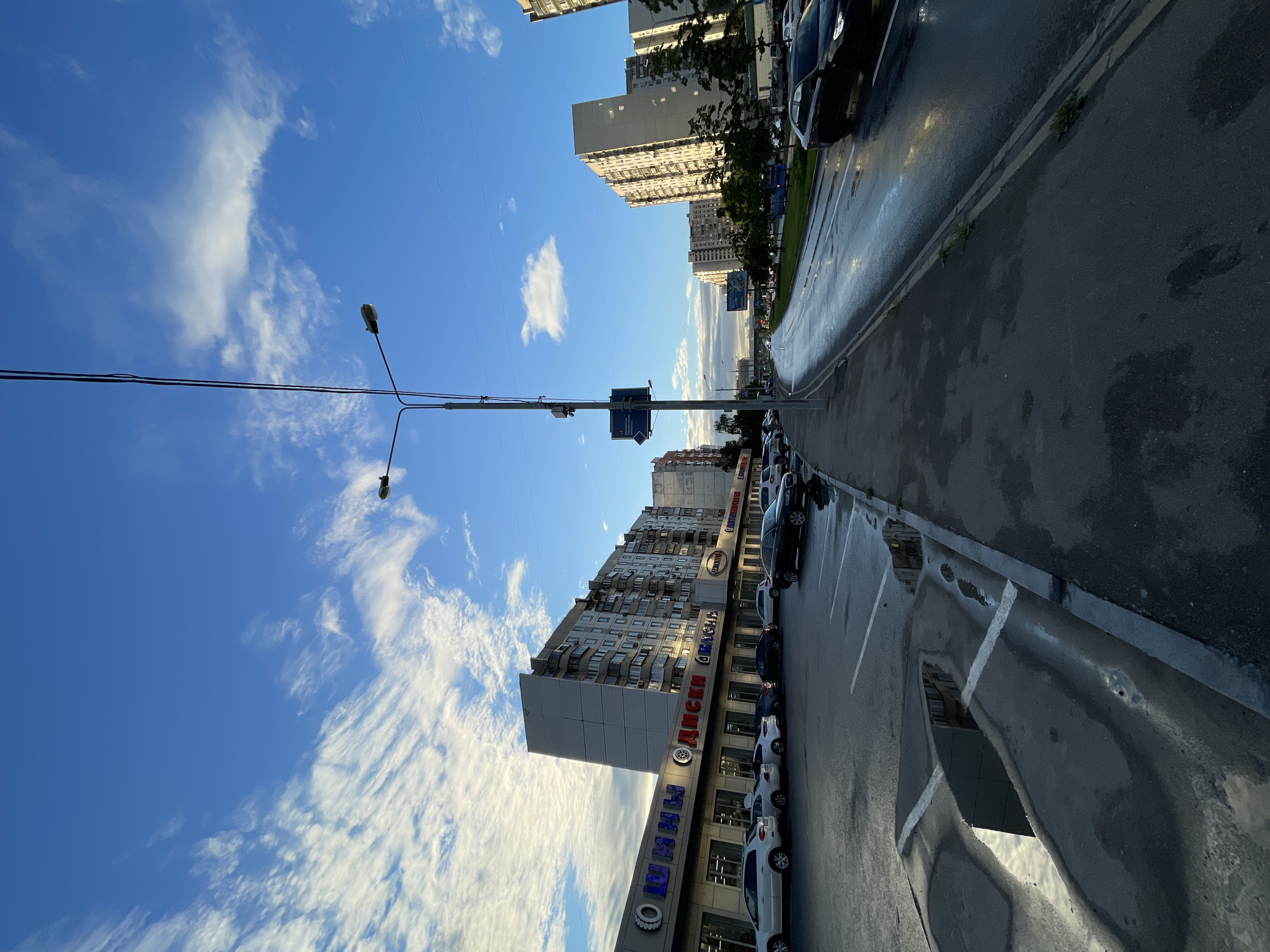This image depicts a street scene in Moscow, Russia. Tall apartment buildings line the right side of a wet asphalt road, reflecting the blue sky with scattered white clouds. The road appears to have recently rained, with visible puddles and reflections of the buildings and streetlights. Parked cars are visible along the curb, in front of a building with a prominent sign that reads "ШИНЫ ДИСКИ" (Tires Disks) in large blue letters with a tire symbol. This suggests a commercial area with automotive-related services. A traffic light is visible on a pole, and power lines stretch across the sky. The lighting suggests it is late afternoon or early evening, with the sun casting long shadows and highlighting the wet surfaces. The overall atmosphere is urban and functional, with a sense of everyday city life.