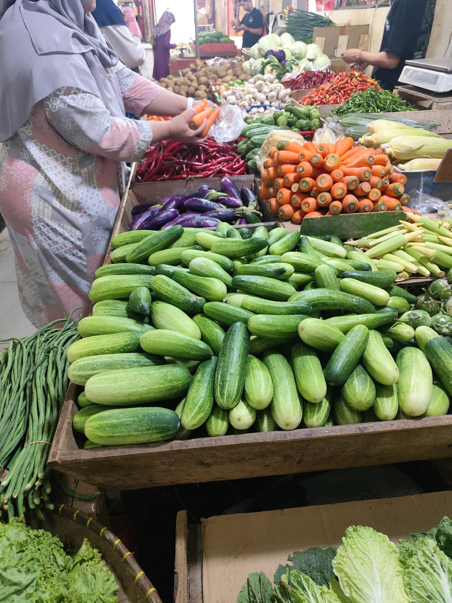This is an eye-level, medium shot of a bustling produce market in Bogor, Indonesia, likely an indoor or covered market given the lighting and stall structures. In the foreground, a large wooden crate overflows with a generous amount of fresh, green cucumbers, some smooth-skinned and others slightly ribbed. To the left of the cucumbers, a bundle of long, dark green beans is tied together with a string. In the middle ground, behind the cucumbers, a person wearing a light purple hijab and a patterned grey and pink garment is sorting through bright orange carrots, holding two in their hands. Behind the carrots, a vibrant pile of red chili peppers is visible. Further back, an assortment of vegetables including eggplants, corn, and cabbages are displayed on stalls. The background is filled with more market stalls and shoppers, suggesting a lively and busy atmosphere. The overall impression is one of abundance and fresh produce, characteristic of Indonesian markets.