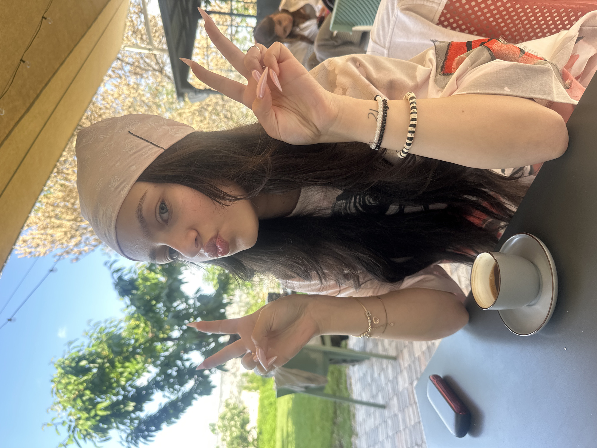 A young woman poses at a table outdoors, making a peace sign with both hands. She is wearing a headscarf and bracelets. A cup of coffee sits on the table next to a red and grey device. Greenery and a blue sky are visible in the background. The location is a cafe in Yerevan, Armenia.