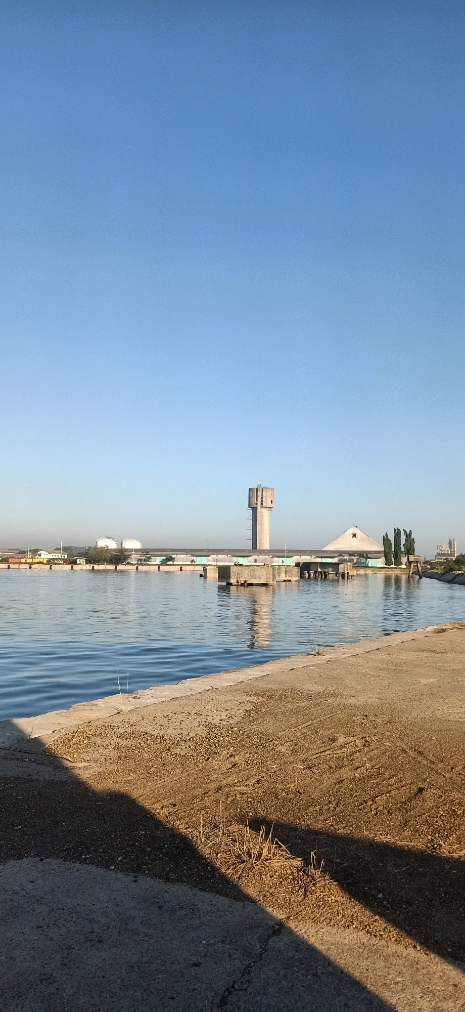 The image depicts an industrial waterfront scene in Keudekruenggeukueh, Indonesia, under a clear blue sky. In the foreground, a concrete embankment lines a body of water. The water is calm with gentle ripples. Beyond the water, a long, low industrial building stretches across the horizon, punctuated by several large storage tanks in the distance. Dominating the mid-ground is a distinctive tall, concrete tower, appearing to be a water tower or industrial chimney. To its right, a large, triangular-shaped structure with a pointed peak is visible. A bridge spans the water in front of the industrial complex. Several trees stand near the triangular structure. In the foreground, dry grass and debris are scattered on the embankment. The overall impression is one of a working port or industrial area.