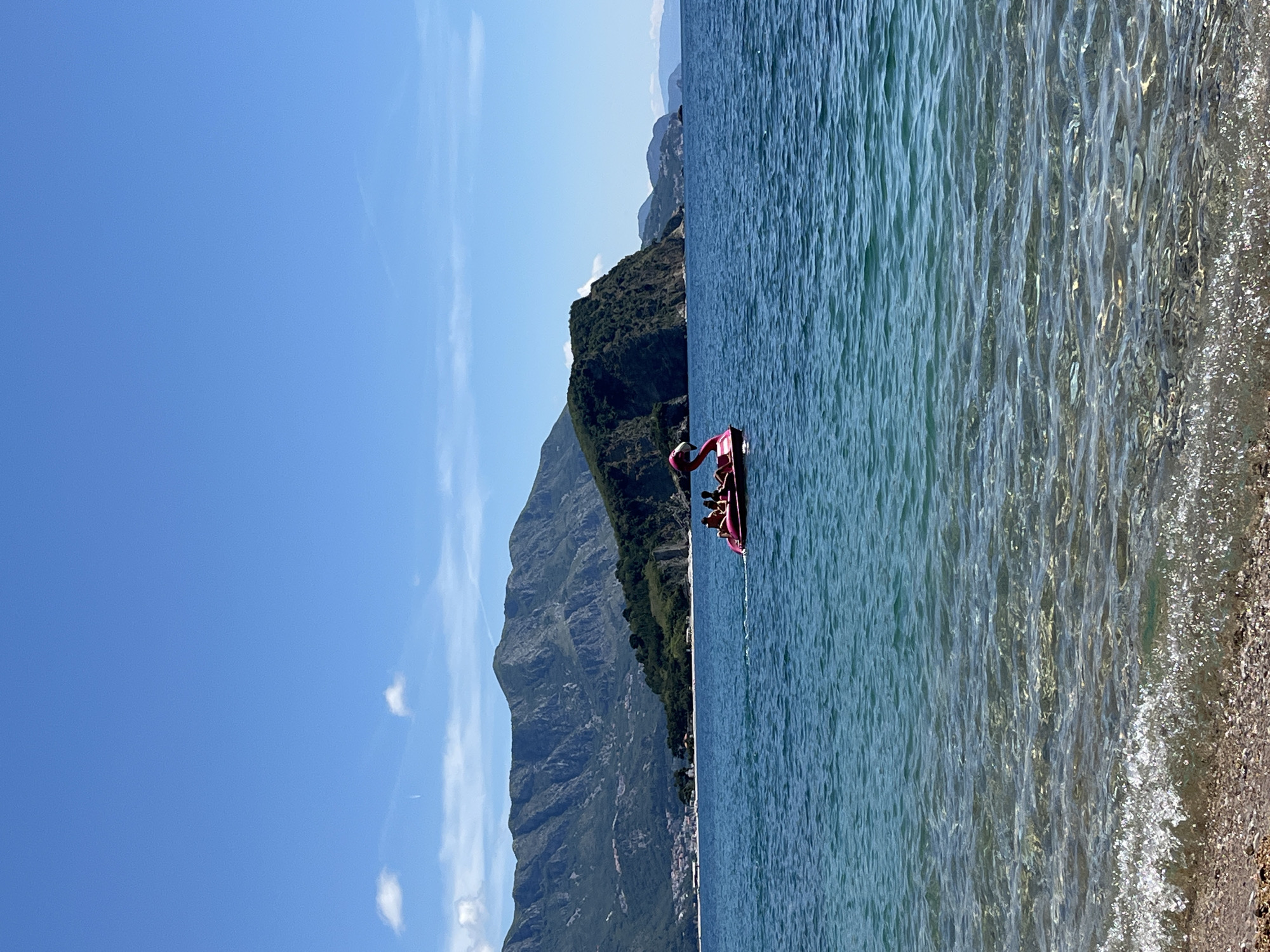 The image shows a coastal scene, likely in Sveti Stefan, Montenegro, given the rocky terrain and clear water. A pink flamingo-shaped paddle boat sits in the water, carrying several people. The water varies in color from turquoise near the shore to darker blue further out. The shoreline is visible on the right, with a mixture of sand and pebbles. A large, mountainous landmass dominates the left side of the image. The sky is blue with a few wispy clouds.