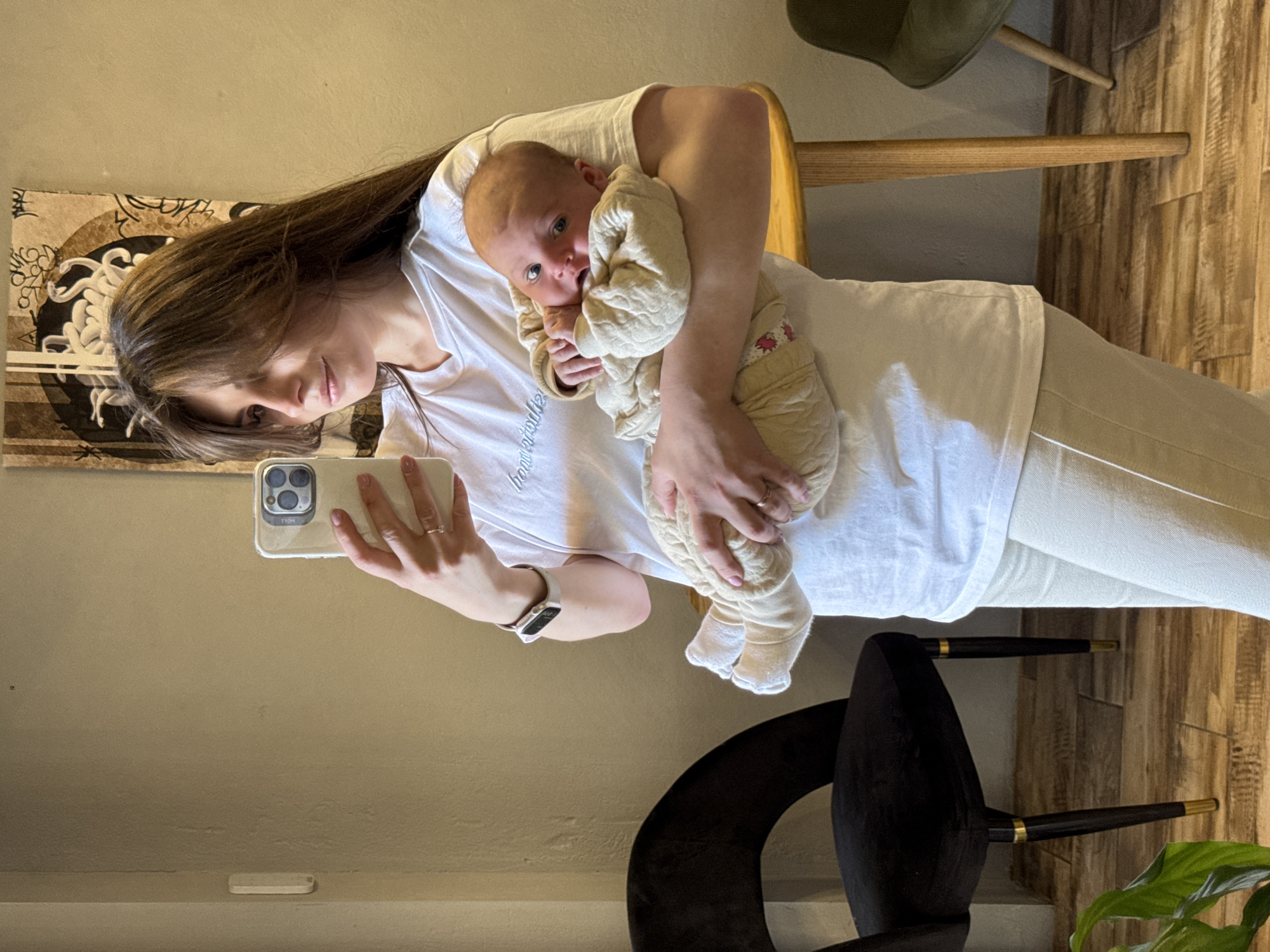 A woman is holding a baby while taking a selfie in what appears to be an indoor setting. The woman has long brown hair and is wearing a white t-shirt and light-colored pants. She is holding a smartphone with both hands, capturing the moment. The baby is wrapped in a light-colored, quilted outfit. The background includes a wall with a decorative piece, a wooden chair, and part of a black chair. The presence of chairs and decorative elements suggests the setting might be a home interior or a cafe. The location is Sofiivska Borshchahivka, Ukraine.