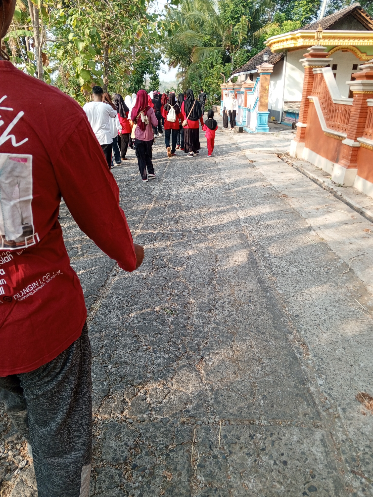 In Lemahbang, Indonesia, a group of people, including men, women, and children, are walking down a paved road. The foreground shows a person from the back, wearing a red long-sleeved shirt with white writing and grey patterned pants. The road has a cracked and worn surface with shadows cast across it from overhead foliage. In the background, on the right side of the road, is a building with an orange and white facade, featuring decorative elements and a tiled roof. There are also trees and other vegetation lining the road and surrounding the buildings. The people are dressed in various attire, with some women wearing headscarves. The overall scene depicts a casual outdoor activity on a sunny day.