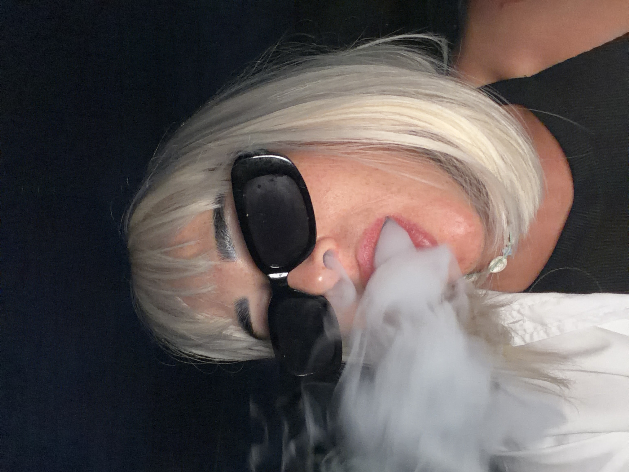 A close-up, eye-level, outdoor shot shows a woman with short, blonde hair and a white wig, wearing large black sunglasses and a black tank top. She is exhaling a large cloud of white smoke from her mouth, which obscures the left side of her face and hair. Her lips are slightly parted, and she is wearing light pink lipstick. A white button-up shirt is visible underneath her tank top on the left side of the frame. A light-colored necklace with a teardrop-shaped pendant is visible around her neck. The background is dark and out of focus, suggesting a nighttime setting. The image does not provide any information about the location being Sochi, Russia.