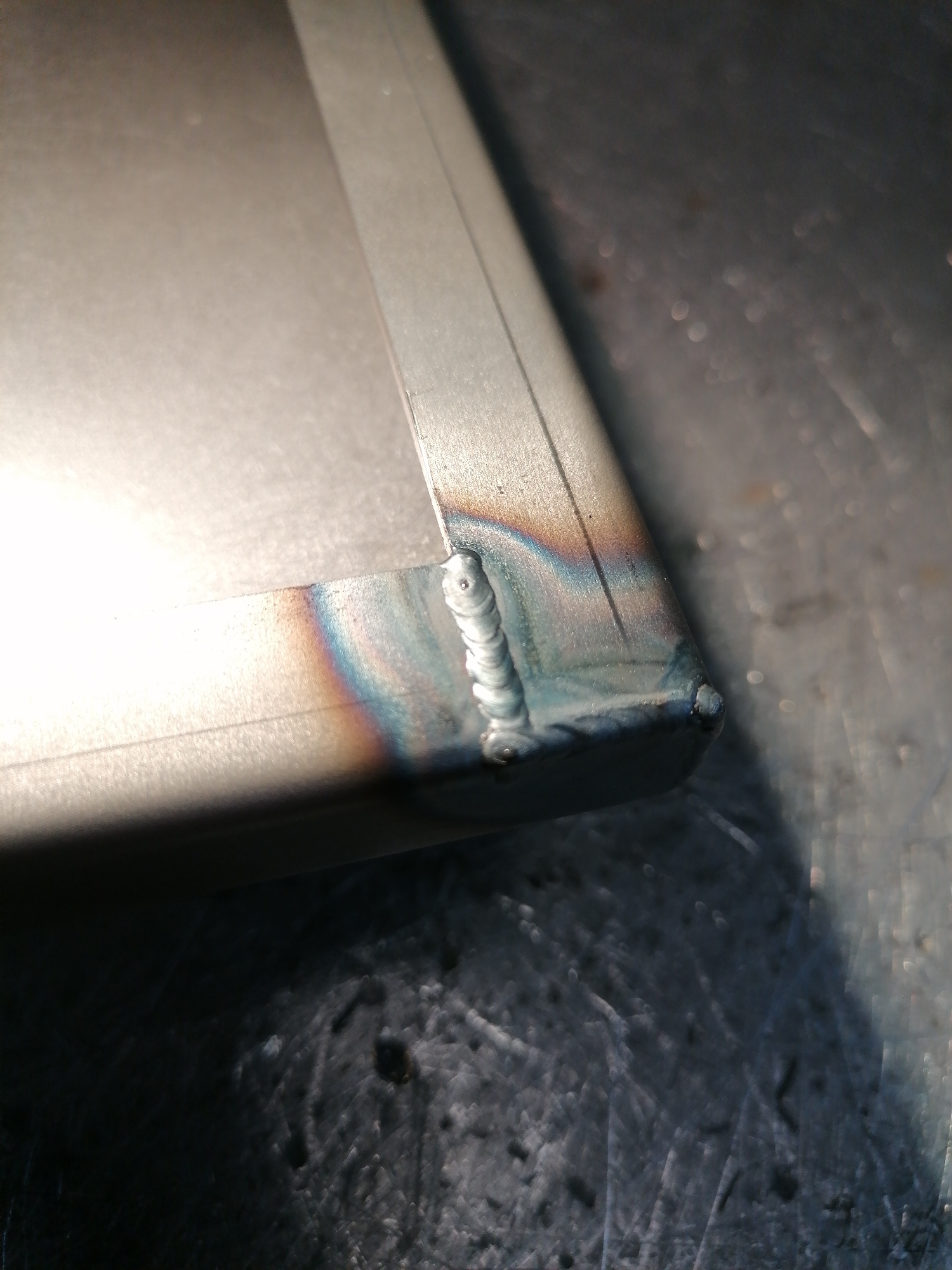The image shows a corner weld on a metal object, likely stainless steel, exhibiting heat tint colors from the welding process. The weld bead is visible, and the surrounding metal shows a rainbow-like discoloration. The metal surface underneath appears to be a work surface with some scratches and imperfections. The scene is indoors, likely a workshop or fabrication area. No people or specific location details are discernible.