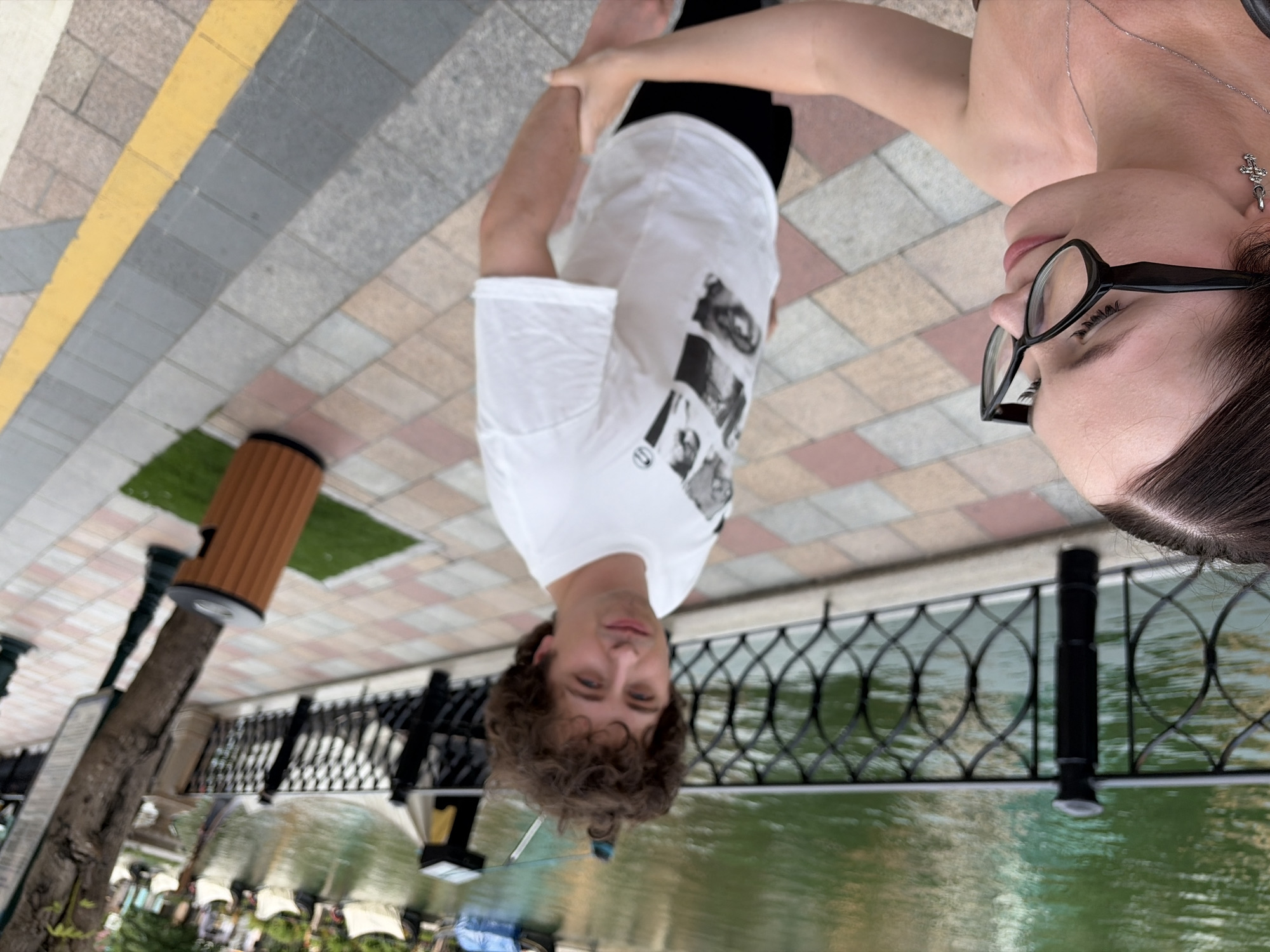 In the foreground, a woman with dark hair, wearing black-rimmed glasses and a silver cross earring, is partially visible from the chest up. She is looking to her left, with her right arm reaching out and her hand resting on the arm of a man. The man, in the midground, has curly brown hair and is wearing a white t-shirt with a graphic print and black shorts. He is looking towards the camera. Behind them, a black metal fence separates a tiled walkway from a body of green water where several gondolas are docked. A tree trunk with some signage is visible to the right of the man. The walkway is paved with a pattern of light brown, cream, and grey tiles, with a yellow line marking an edge on the right side. The environment suggests an outdoor, possibly tourist, location. There is no specific indication of Nghĩa Trụ Commune, Vietnam.