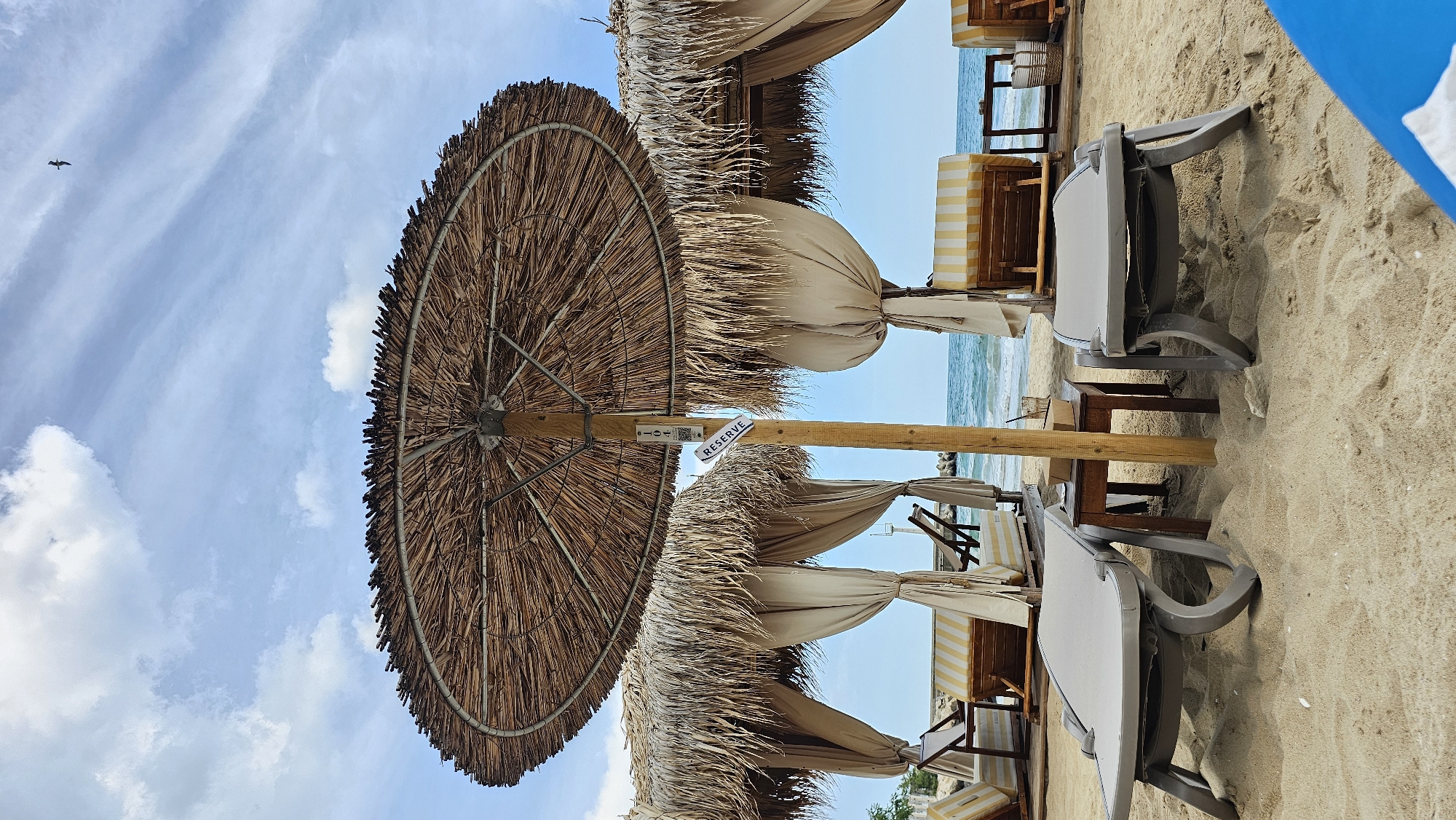 This is a sideways, eye-level shot of a beach scene in Varna, Bulgaria. The foreground is dominated by a large, thatched umbrella, its straw fronds fanning out to the left. A wooden pole supports the umbrella and a sign that reads "RESERVE". To the right of the umbrella, several lounge chairs with beige cushions are arranged on the sand. Behind the chairs, a row of cabanas with striped yellow and white awnings and draped beige fabric line the beach.
The background features the calm, blue sea meeting a bright, light blue sky with scattered white clouds. A single bird is visible in flight in the upper left portion of the frame. The sand is light brown and appears undisturbed, suggesting a peaceful atmosphere. The lighting suggests it is daytime, likely mid-morning or afternoon, with clear weather. The scene evokes a relaxed, holiday mood, with the "RESERVE" sign indicating a commercial establishment preparing for or already serving guests.