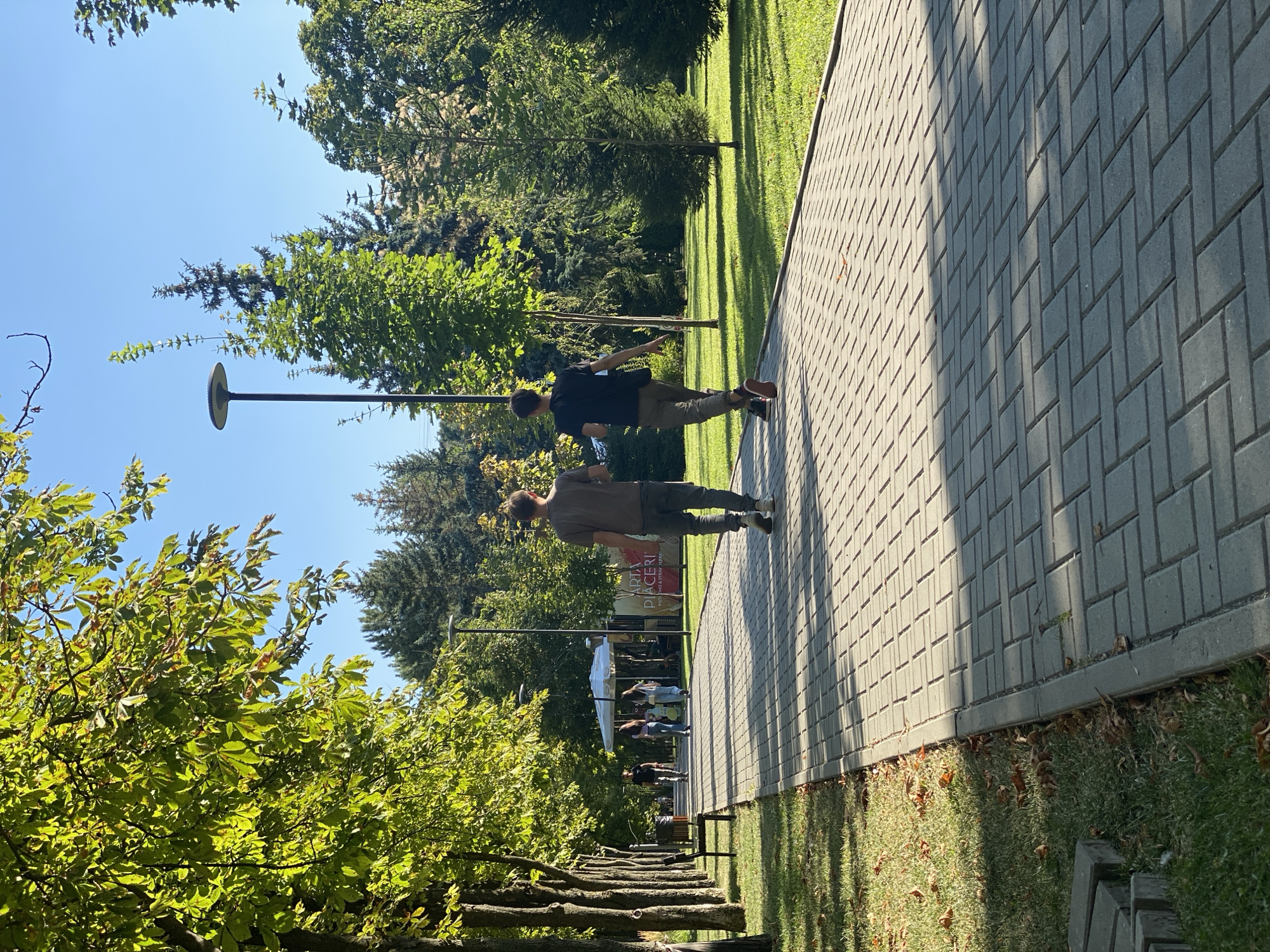 The image depicts a sunny day in a park with a paved walkway. Two young men are walking away from the camera on the walkway. The man in front is wearing a grey t-shirt and dark pants. The man behind him is wearing a black t-shirt and grey pants with red sneakers. In the background, a white umbrella structure is visible, along with other people walking on the path. Trees with green leaves line the walkway on the left side, and a grassy area with scattered fallen leaves is on the right. The sky is clear and blue. The location is likely Dendrarium Park in Chișinău, Moldova, given the style of the walkway and the abundance of trees and greenery.