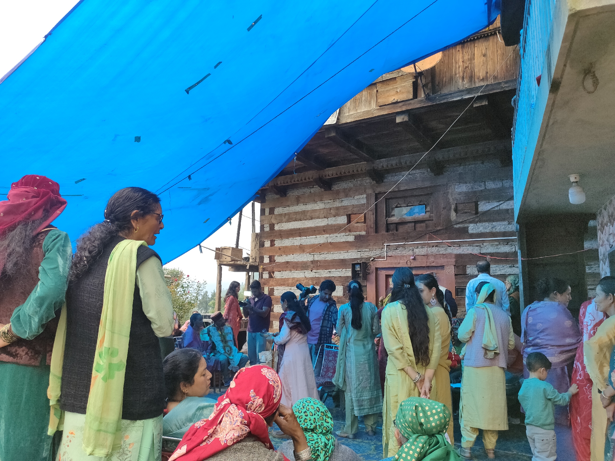In Damadi, India, a group of people are gathered outdoors under a large blue tarp. The setting appears to be a village or rural area, with a rustic wooden building in the background. The people are dressed in traditional Indian attire, with vibrant colors and patterns visible on their clothing and headscarves. Some individuals are talking, while others are observing. A man in the center appears to be holding a camera or video recorder, suggesting a possible event or gathering. The atmosphere seems to be festive and communal.