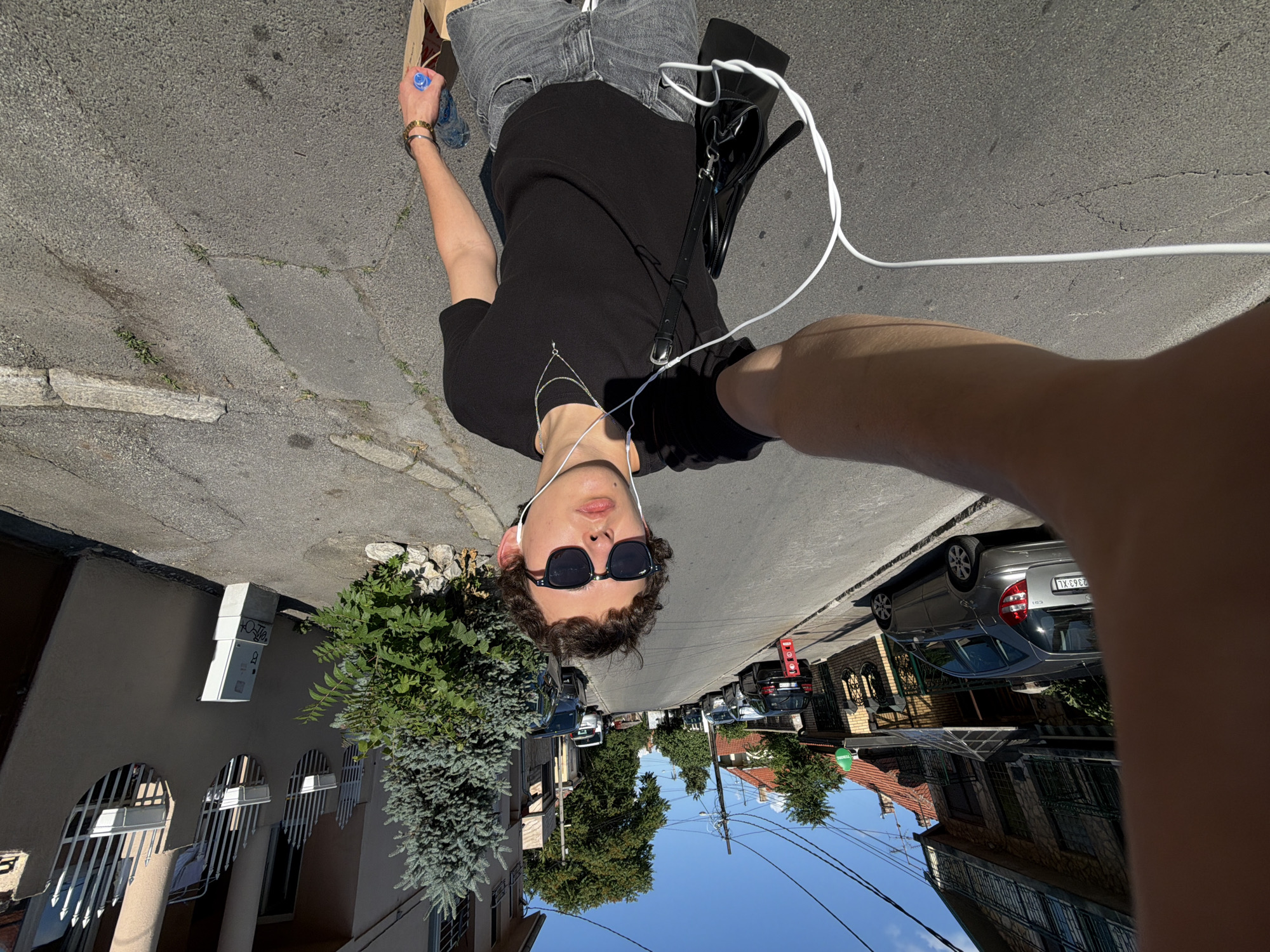 The image depicts a young male taking a selfie on a residential street in Palilula Urban Municipality, Serbia, on a clear, sunny day. The male, positioned in the foreground, has short, curly brown hair and wears dark sunglasses, white wired earphones, a black short-sleeved t-shirt, gray denim shorts, a silver chain necklace, and a gold-colored bracelet on his right wrist. He holds a black shoulder bag on his left side and, with his right hand, a blue-capped plastic water bottle and a light brown paper bag. He extends his left arm to hold the camera, capturing his reflection and the street scene behind him.
The street is paved with asphalt, showing some cracks and repairs, and slopes gently uphill away from the viewer. On the left side of the street, partially obscured by the male's arm, a silver Mercedes-Benz A-Class hatchback is parked, displaying a Serbian license plate with "2363-XL" visible. Behind it, a multi-story building with a stone-patterned facade, dark-framed windows, and ornate railings is visible, along with glimpses of red-tiled roofs further down the street. A small green sign with white lettering is affixed to a building facade. Overhead power lines crisscross against the bright blue sky.
On the right side of the street, a multi-story building with a light beige facade features a distinctive wavy design, incorporating white vertical metal railings and white planter boxes. Against this building, a white utility box rests on a concrete base. A prominent blue spruce tree, with smaller green shrubs at its base surrounded by rocks, stands next to the sidewalk. The sidewalks are concrete slabs, with patches of grass growing in the cracks, separated from the road by concrete curbs. Several other vehicles, including a black car and a white van, are parked further up the street. The bright sunlight casts distinct shadows, indicating mid-day.