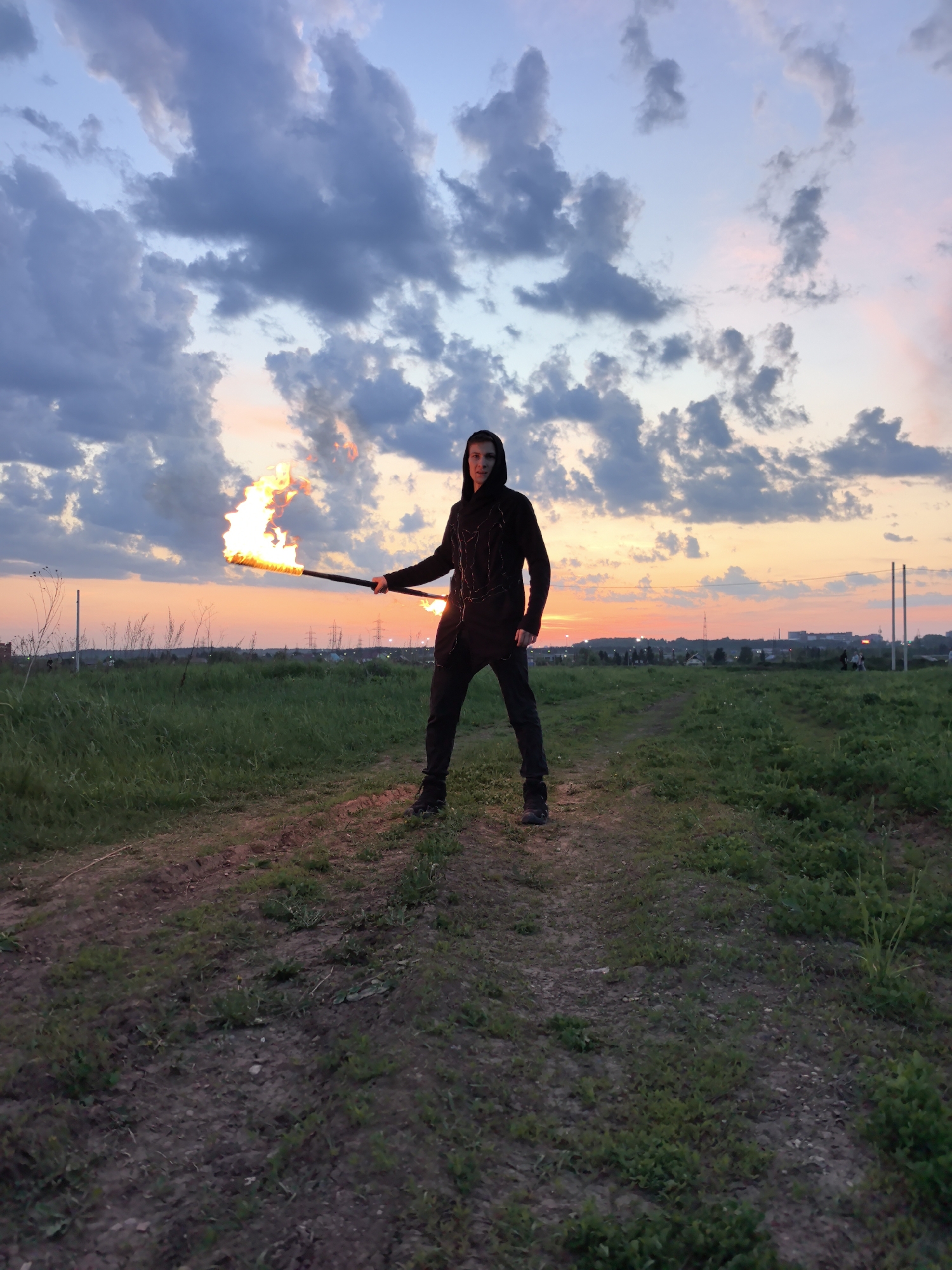 The image shows a person standing in a field on a dirt path. The person is holding a flaming torch in their hands. The person is wearing a dark hooded jacket and dark pants. The sky behind the person is cloudy with a sunset in the background. In the distance, there are buildings and power lines. The image was taken in Фролы, Russia.