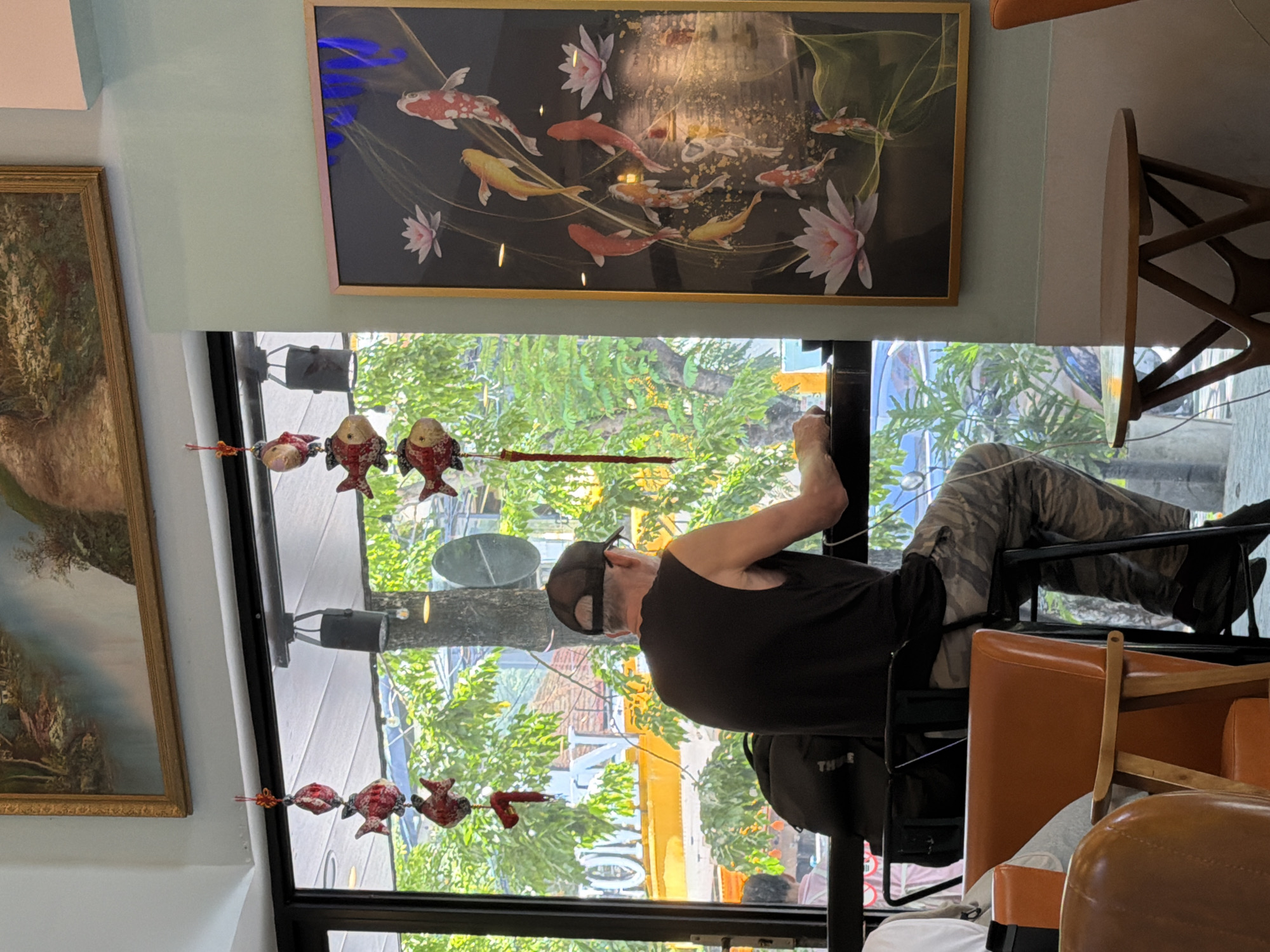 A man is cleaning a window in a building in Đà Nẵng, Vietnam. The man is wearing a black tank top, camouflage pants, and a baseball cap. He is using a squeegee to clean the window. There are several paintings on the wall, including a painting of koi fish. There are also several fish-shaped decorations hanging from the window. The background shows a street scene with trees and buildings. There is a chair and a table visible on the right side of the frame.