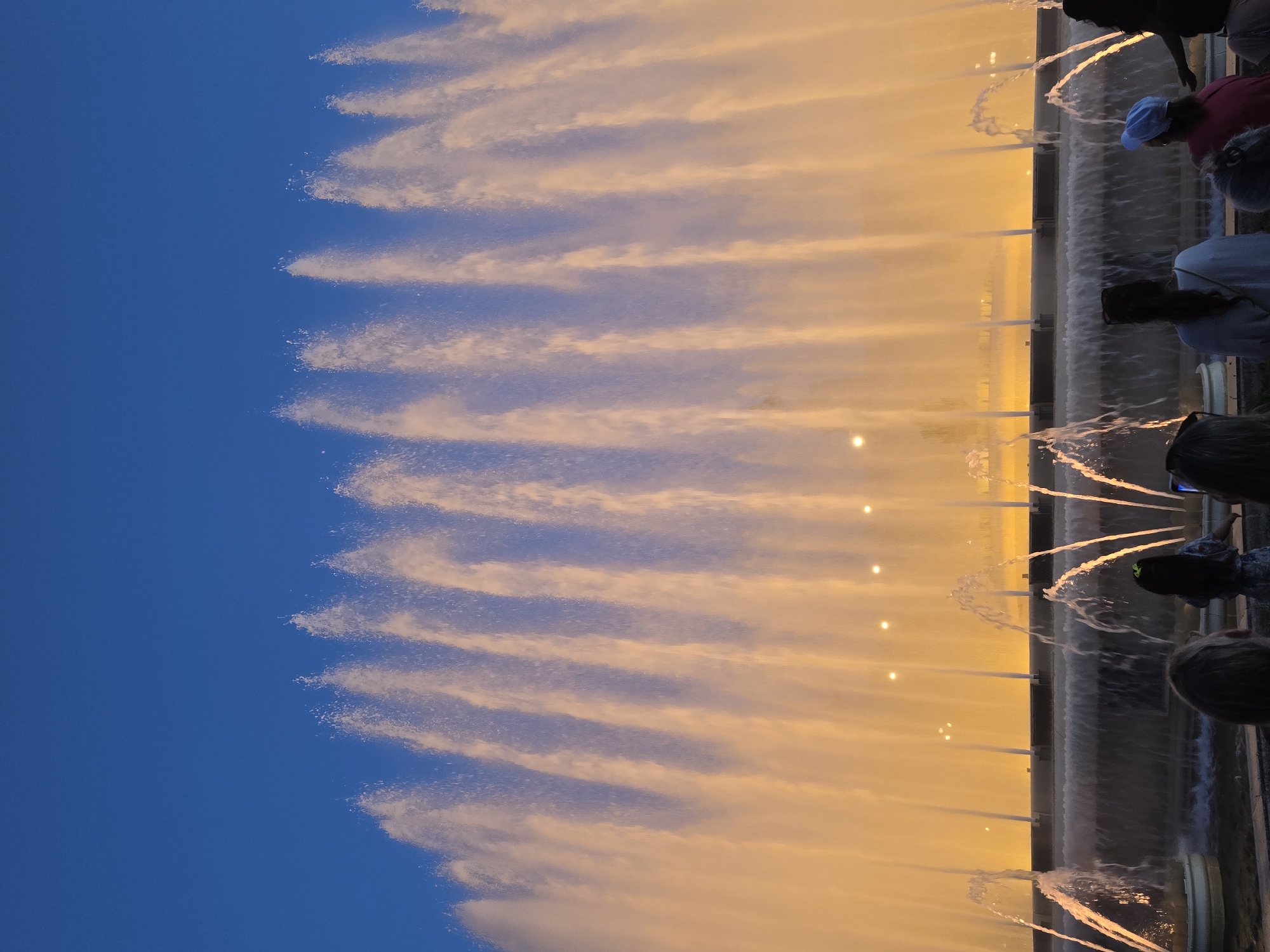 The image depicts the Magic Fountain of Montjuïc in Barcelona, Spain, at dusk. The fountain is illuminated with warm yellow light, casting a glow on the cascading water jets that reach high into the twilight sky. The sky is a deep, clear blue, transitioning to a lighter hue near the horizon. In the foreground, a group of silhouetted figures are gathered, facing the fountain and seemingly observing the spectacle. The people are mostly out of focus, with only their outlines visible, suggesting they are standing some distance away. The fountain itself is a tiered structure, with multiple levels of water flow and spray. Small lights are visible within the water displays and on the fountain's structure, contributing to the overall illuminated effect. The scene conveys a sense of awe and wonder, with the vibrant lights and water creating a dynamic and captivating display.