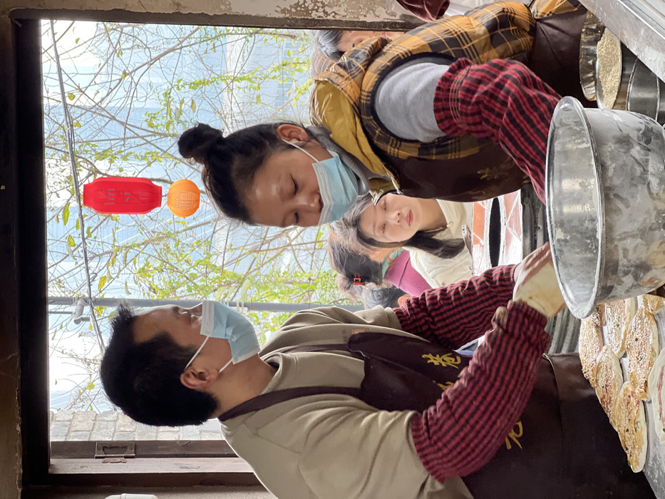 Several people are positioned around what appears to be a food preparation area. One person, wearing a blue face mask and a brown apron over a beige shirt and red striped sleeves, is looking down at a cooking surface with multiple circular food items. Another person wearing a blue face mask, a yellow plaid vest, and red sleeves is also looking down. Two red and orange lanterns hang outside the window next to bare tree branches.