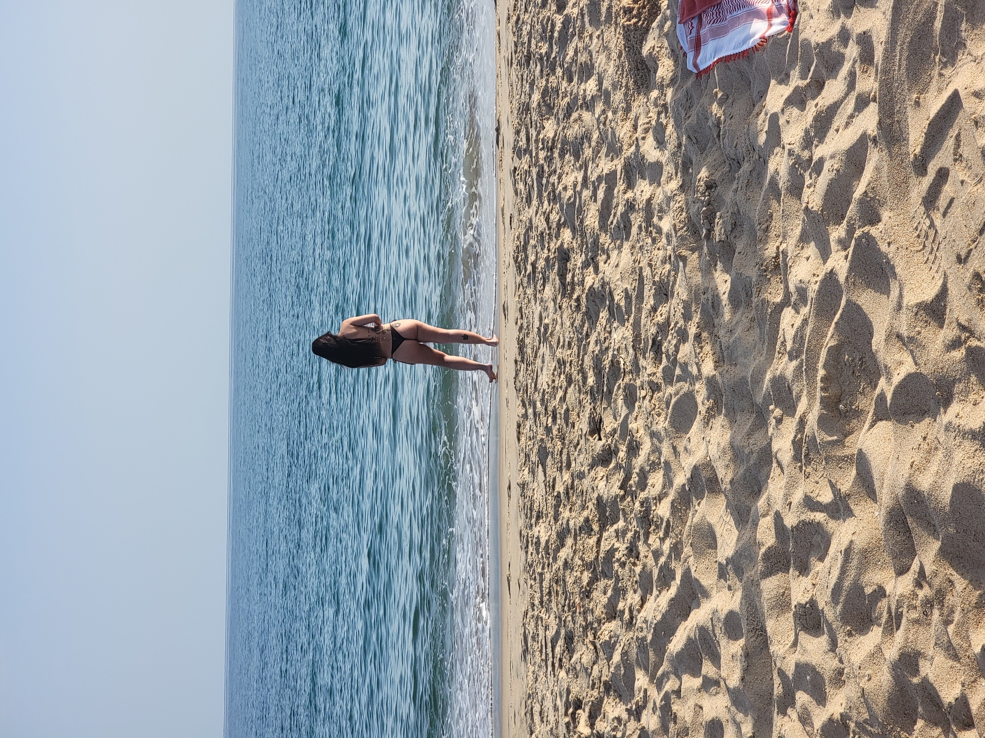 A person stands on a sandy beach, facing the ocean. The person has long, dark hair and is wearing a black bikini. Their feet are in the shallow water at the edge of the sea, where small waves are breaking. The sand in the foreground is textured with ripples and shadows from the sunlight. In the upper right corner of the frame, a folded towel with red and white stripes is visible on the sand. The ocean is a deep blue, and the sky is a clear, light blue, meeting the horizon in a thin line. This scene is located on a beach in Quarteira, Portugal.