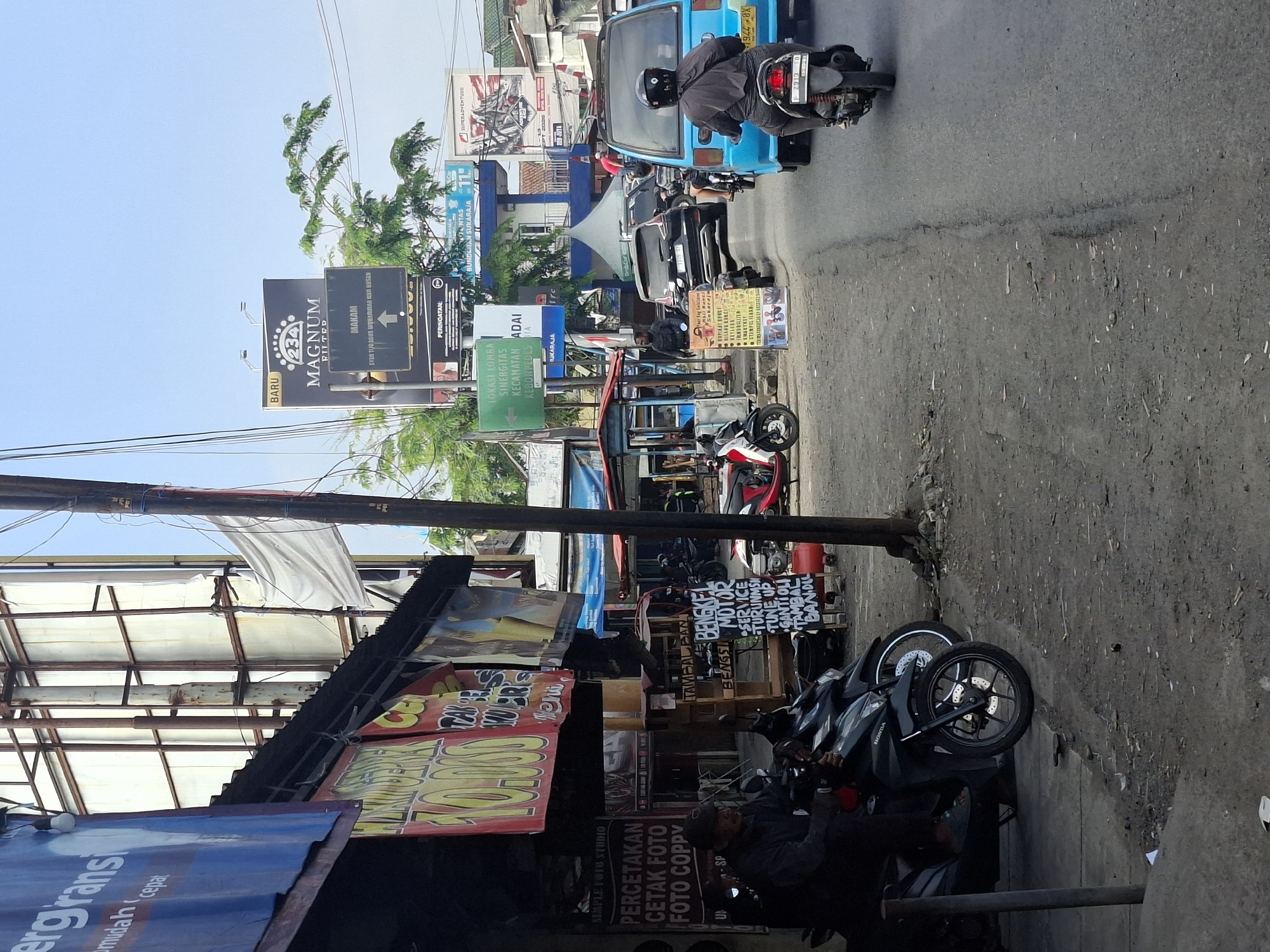 The image depicts a street scene in Sukaraja, Indonesia, characterized by commercial activity and traffic. In the foreground, several motorcycles are parked, with one rider seated on a dark-colored Honda motorcycle. Adjacent to the motorcycles is a storefront with various banners and signs advertising services such as "PERCETAKAN CETAK FOTO FOTO COPY" (Printing, Photo Printing, Photo Copy). Another sign indicates "BENGKEL MOTOR" (Motorcycle Workshop) offering services like "SERVICE TURUN MESIN" (Engine Overhaul Service), "TUNE UP," "GANTI OLI" (Oil Change), and "TAMBAL BAN" (Tire Patching).
Further down the street, traffic is visible, including a blue van and several other vehicles. A man in a black shirt and helmet is riding a motorcycle in the blue van's lane. Billboards are present in the background, one of which advertises "MAGNUM FILTER" and mentions "MAKAM SYEK TUGUS ABDURRAHMAN BIN HUSEIN" (Tomb of Syek Tugus Abdurrahman bin Husein). Other signs are in Indonesian, suggesting the local context. The environment appears to be a busy urban area with a mix of buildings and infrastructure. The sky is clear and bright, indicating daylight.
