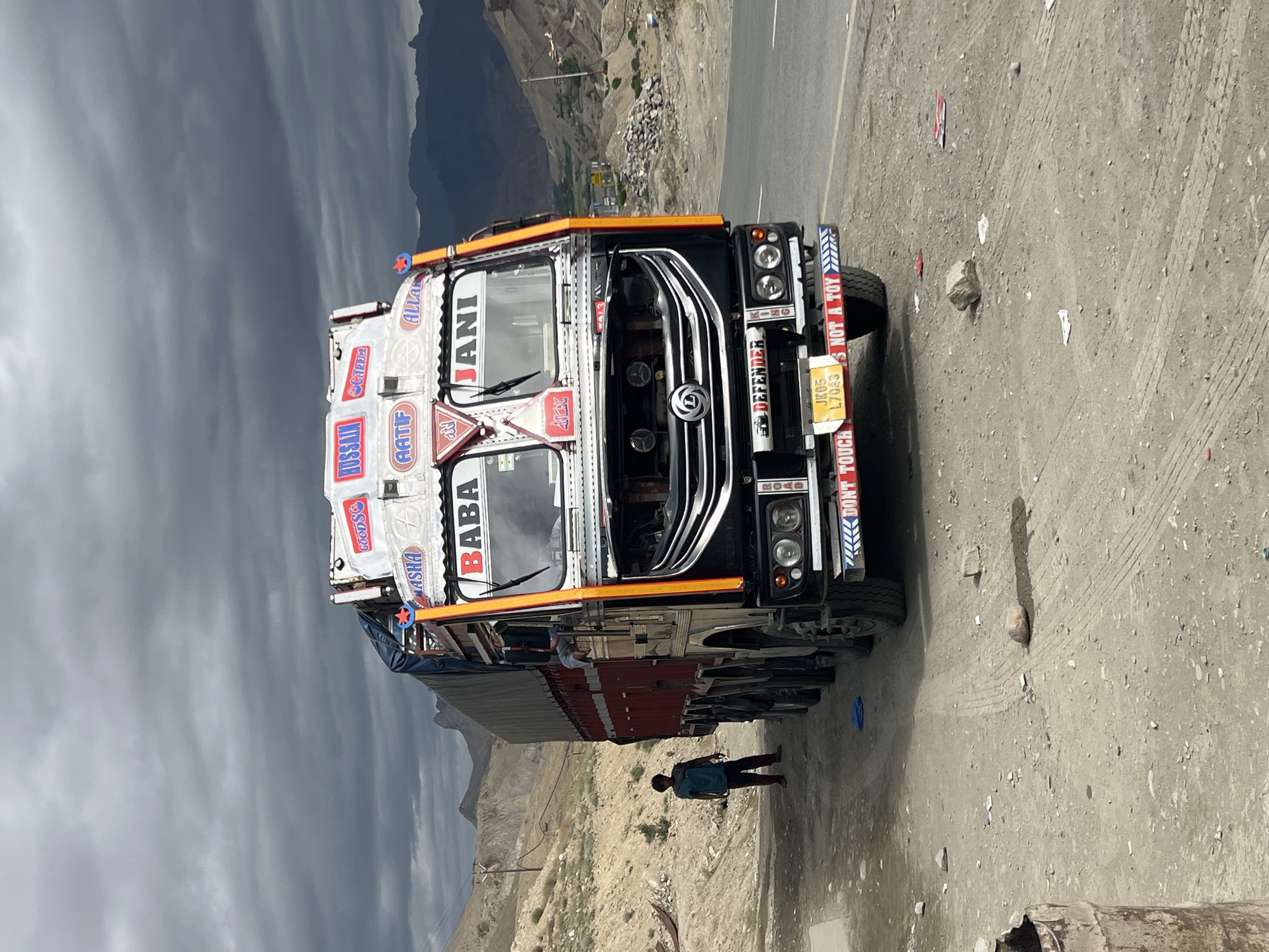 A heavily decorated, black and red truck is parked on the side of a dusty road in a mountainous, arid region of India. The truck has numerous decals and text on its exterior, including "BABA JANI" on the windshield, and "HUSSAIN" and "GOODSG" on the top. A young boy wearing a teal shirt and dark pants stands to the left of the truck. The sky is overcast with dark clouds. The surrounding landscape consists of barren, rocky hills.