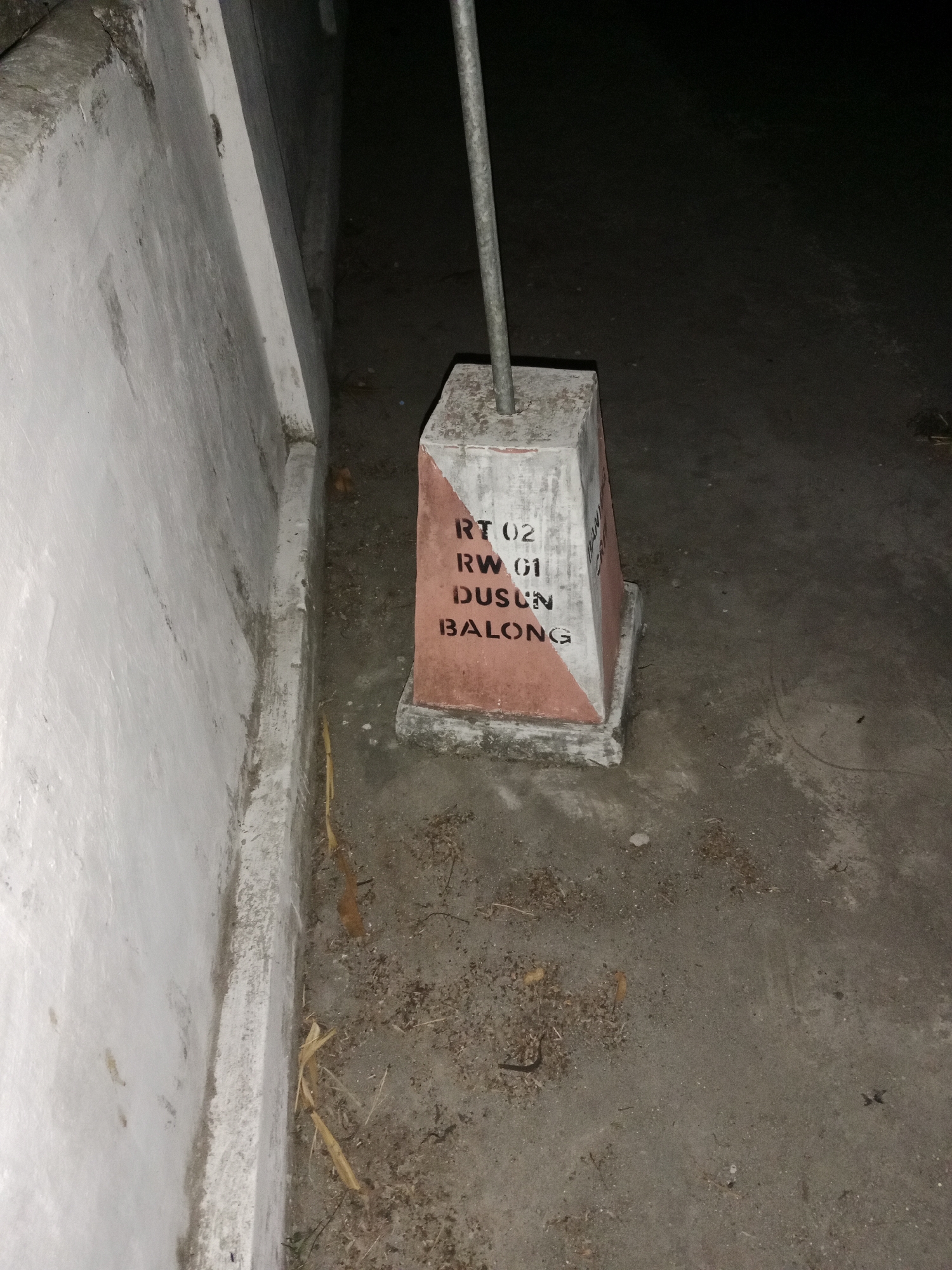 The image captures a concrete marker post with black lettering and red and white diagonal markings. The text on the post reads "RT 02 RW 01 DUSUN BALONG". The marker is situated outdoors on a sandy or gravelly surface. To the left of the marker, there is a light-colored, textured wall, possibly made of concrete or stucco, which appears to be part of a building or structure. The background is dark and out of focus, suggesting it is nighttime or a poorly lit area. There are scattered dried leaves and debris on the ground around the marker. A thin metal pole extends vertically from the top of the marker post, disappearing out of the frame. The overall scene is a street-level view of a marker identifying a specific residential area (RT/RW) and a hamlet (Dusun) in Ringinrejo, Indonesia.