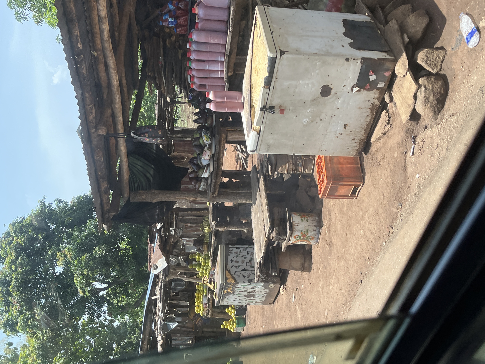 This image captures an outdoor market stall or roadside shop in Karshi, Nigeria, under bright daylight. The setting features a rustic structure built from wooden poles and corrugated metal sheets, providing shade over various goods displayed on wooden tables and shelves.
Key subjects include:
- **Produce:** Several bunches of green bananas or plantains hang from the stall's wooden framework. Piles of green, round fruits, possibly limes or small green mangoes, are arranged on a low table. Another wooden crate holds small orange items, likely tomatoes or oranges.
- **Packaged Goods:** Rows of clear plastic bottles containing a pinkish-red liquid, consistent with palm oil, are neatly displayed on an upper wooden shelf.
- **Equipment:** A large, weathered white chest freezer or cooler, appearing old and possibly non-functional, is prominent near the center, with its insulation partially exposed. It has some indistinct stickers on its side. An old, decorated metal bucket or container sits on the ground near one of the stalls.
- **Environment:** The ground is dirt, interspersed with several rough stones. Large green tree foliage is visible in the background and to the left, indicating a lush natural environment. The sky is clear blue with minimal clouds, suggesting sunny weather.
No people are clearly visible, but the setup implies active commerce. A white flip-flop and a small dark object are visible on the dirt ground. No legible text is discernible on any of the items. The scene portrays a functional, open-air retail environment typical of the region.