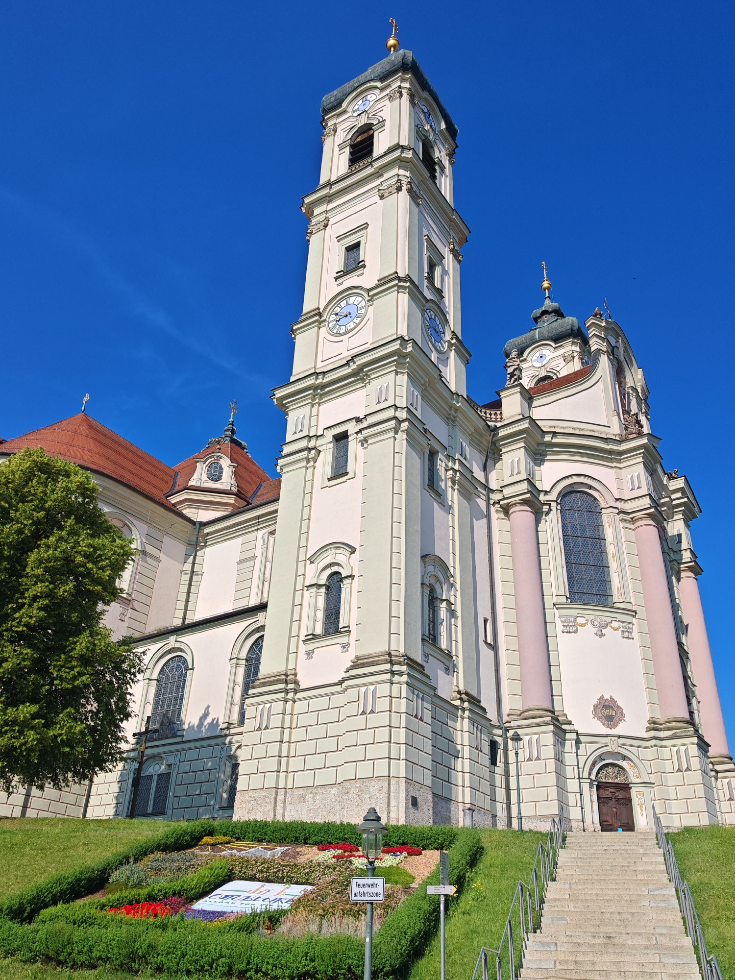 The image captures the Basilica of St. Alexander and St. Thekla in Ottobeuren, Germany, on a clear, sunny day. The imposing Baroque church dominates the frame with its cream-colored facade, adorned with light green and pink architectural details. A tall clock tower, topped with a golden cross, rises majestically against a vibrant blue sky. Multiple clock faces are visible on the tower.
In the foreground, a well-maintained grassy slope leads up to the church. A colorful flowerbed, arranged in a patterned design, decorates the lower part of the slope. In front of the flowerbed, a sign with German text, possibly indicating a fire department access zone, is visible. Stone steps with metal railings ascend towards a wooden entrance door of the basilica. A large green tree stands to the left of the church. The overall scene depicts a serene and picturesque view of a significant religious landmark.