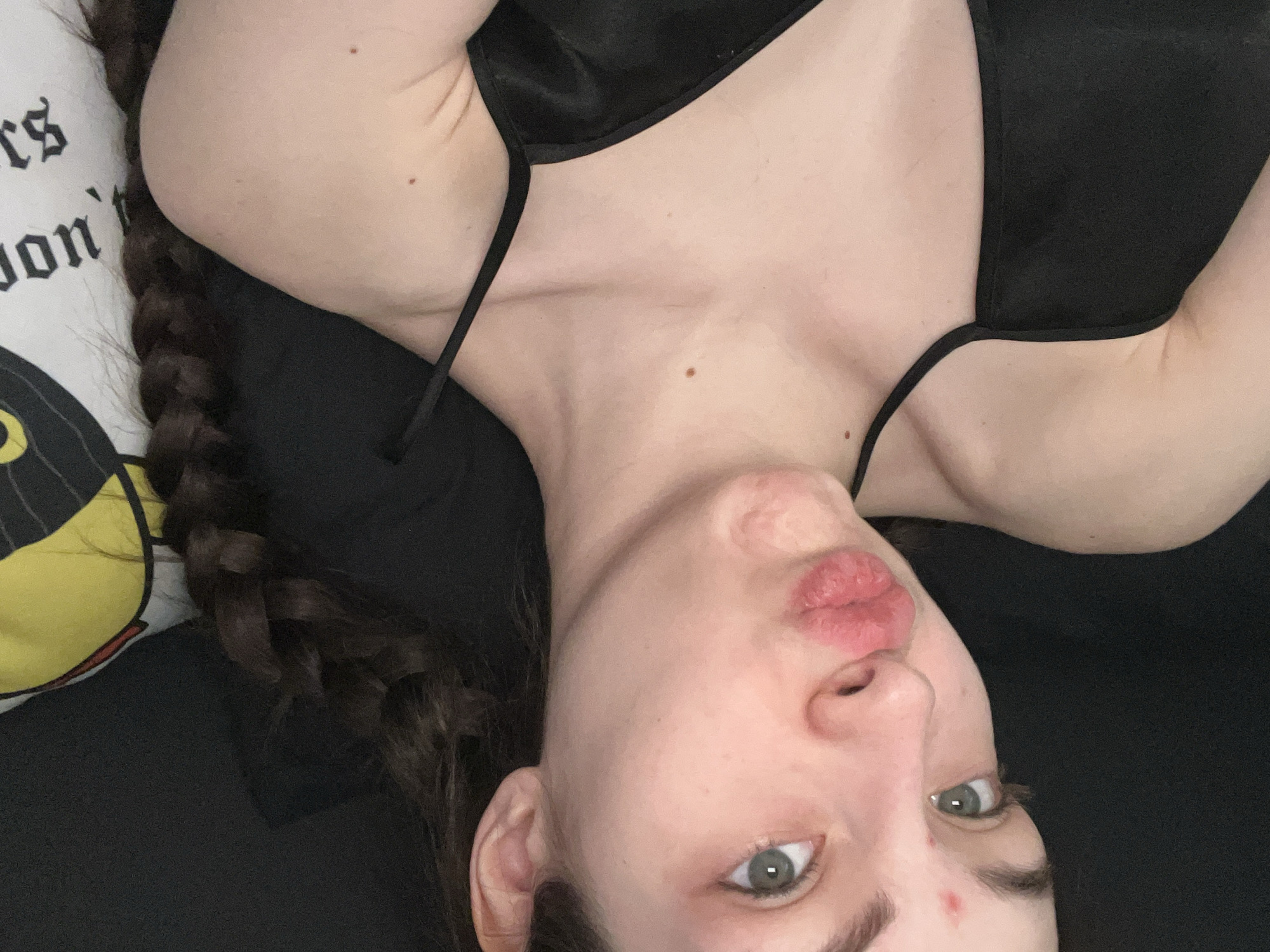 A person is lying down, facing the camera. They have a braid in their hair and are wearing a black top. The person's lips are pursed. A pillow with text is visible next to their head. The image does not contain enough information to determine the location.
