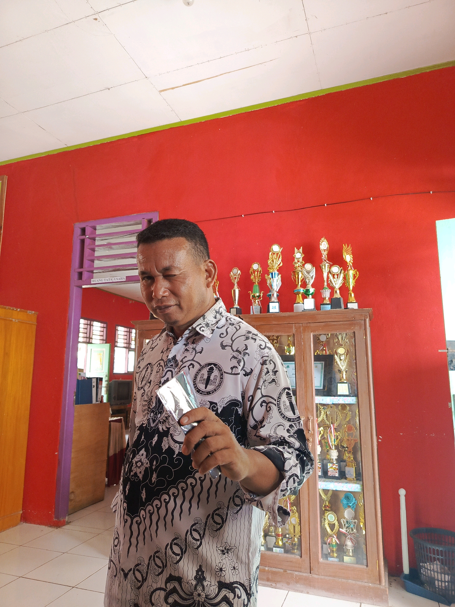 A man wearing a black and white patterned shirt is standing in a room with red walls and a white ceiling. He is holding a silver object, possibly a phone or a card, in his right hand and winking at the camera. Behind him, there is a wooden cabinet filled with trophies. The room appears to be in a school or a community center, given the trophies and the sign above a doorway that reads "RUANG TATA USAHA" (Office Administration Room). The man is the central subject of the image. The background includes furniture, doors, and windows. The lighting suggests an indoor setting with overhead lights. The man's expression is playful and engaging.