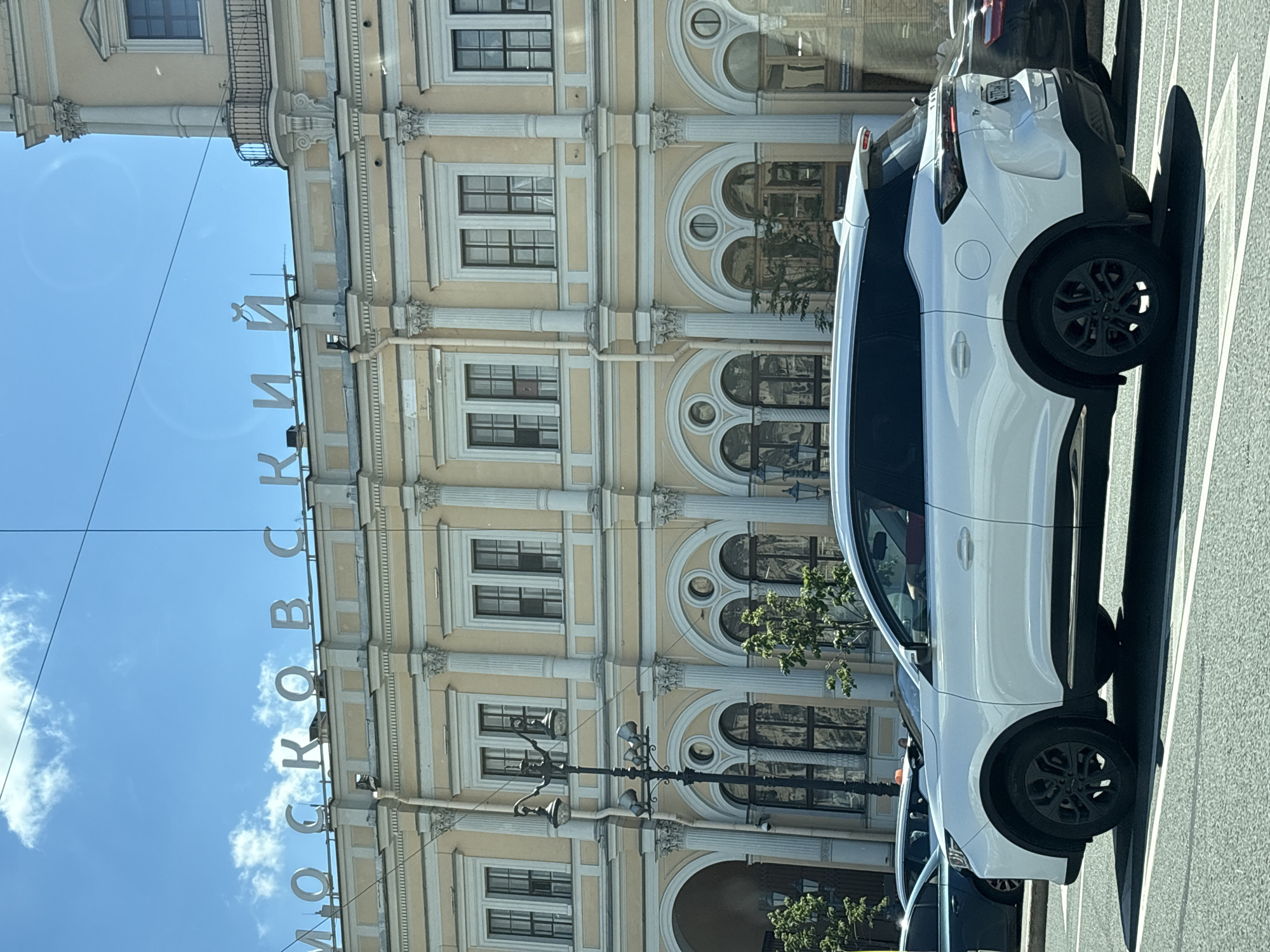 The image shows a street scene in Saint Petersburg, Russia, with a large, light-yellow building in the background. White Cyrillic lettering, spelling "МОСКОВСКИЙ", is mounted on the roofline of the building. The building features classical architectural elements, including columns, arched windows on the lower level, and rectangular windows on the upper level, framed by decorative molding. A street lamp with multiple lights stands in front of the building. In the foreground, a white SUV is parked on the street, with other vehicles partially visible. The sky is blue with scattered white clouds, and overhead power lines are visible.