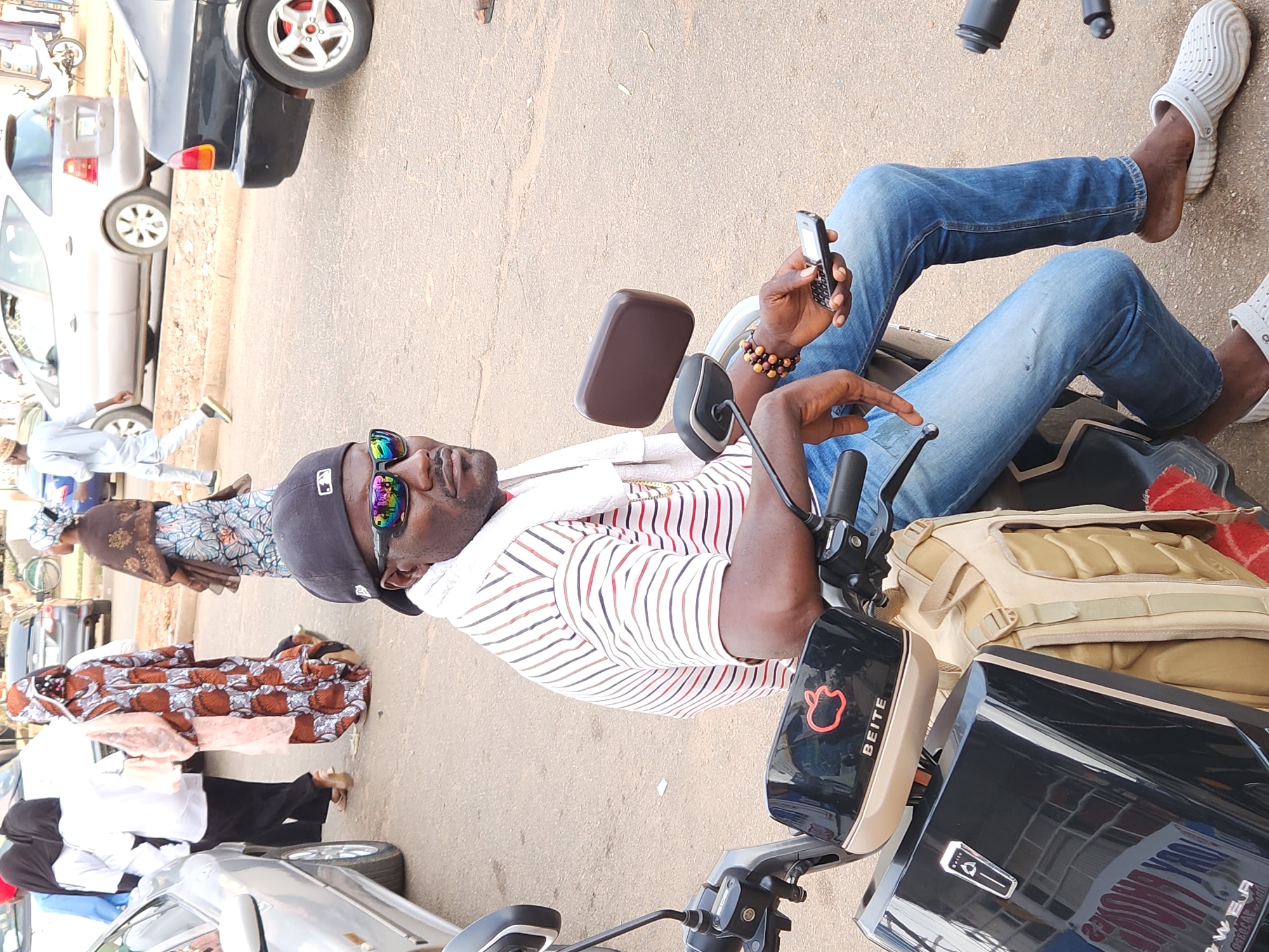 A man sitting on a motorcycle on a paved street in Unknown, Unknown. The man is wearing blue jeans, a striped shirt, a black baseball cap, and sunglasses. He is holding a cell phone in his left hand. The motorcycle is black with a tan side bag. There are several people walking in the background and several cars parked on the side of the road.