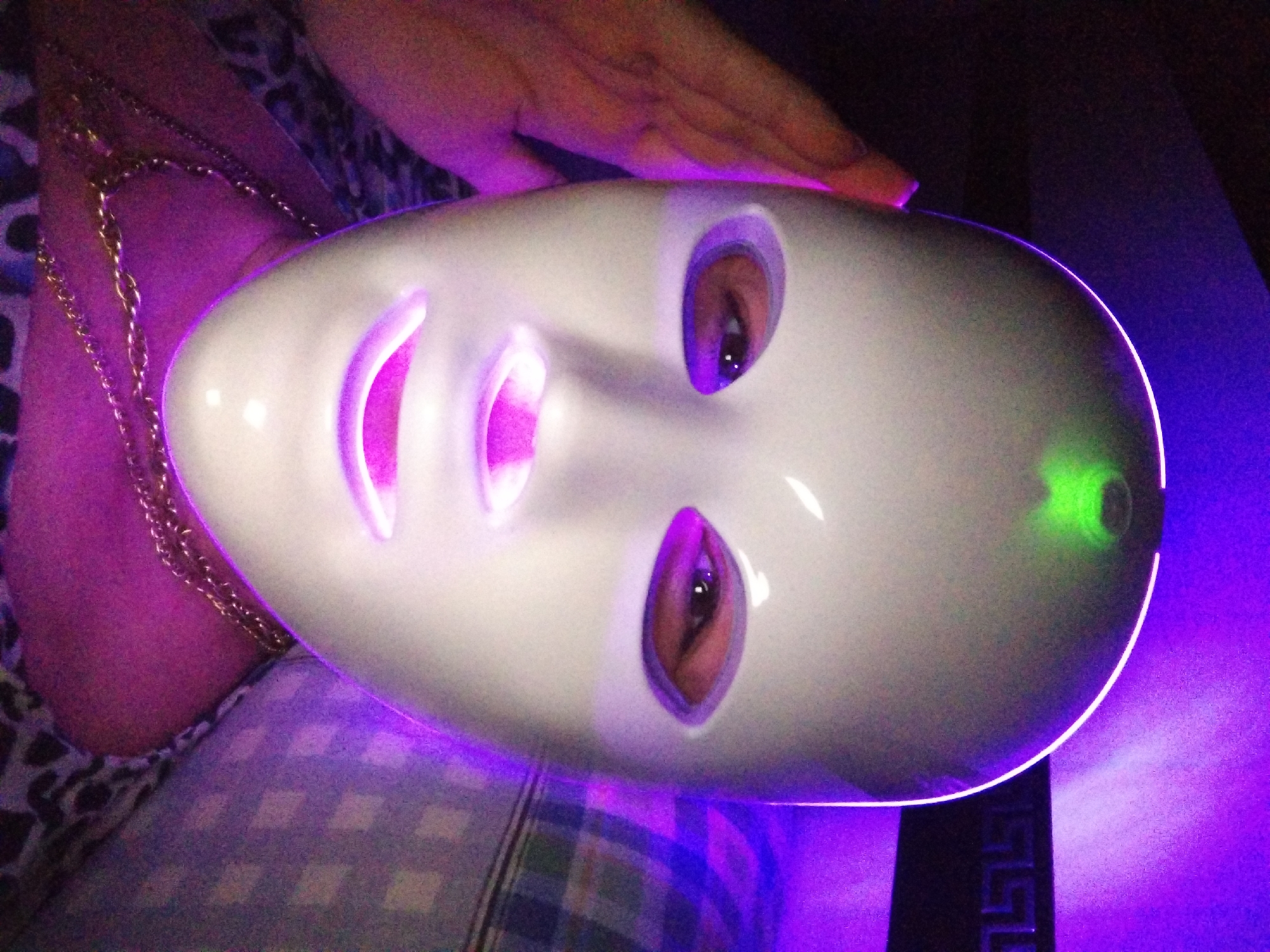 An individual is wearing a white LED facial mask, which is illuminated with purple and green light. The mask covers the majority of their face, with openings for the eyes and mouth. A hand is visible on the left side of the mask, near the temple, and appears to be holding it in place. The individual is wearing a gold chain necklace. The background is dark, with purple light illuminating the right side of the image, suggesting an indoor environment. A patterned surface, possibly a blanket or pillow, is visible on the right. There is no indication of the city of Stara Zagora, Bulgaria, in the image.