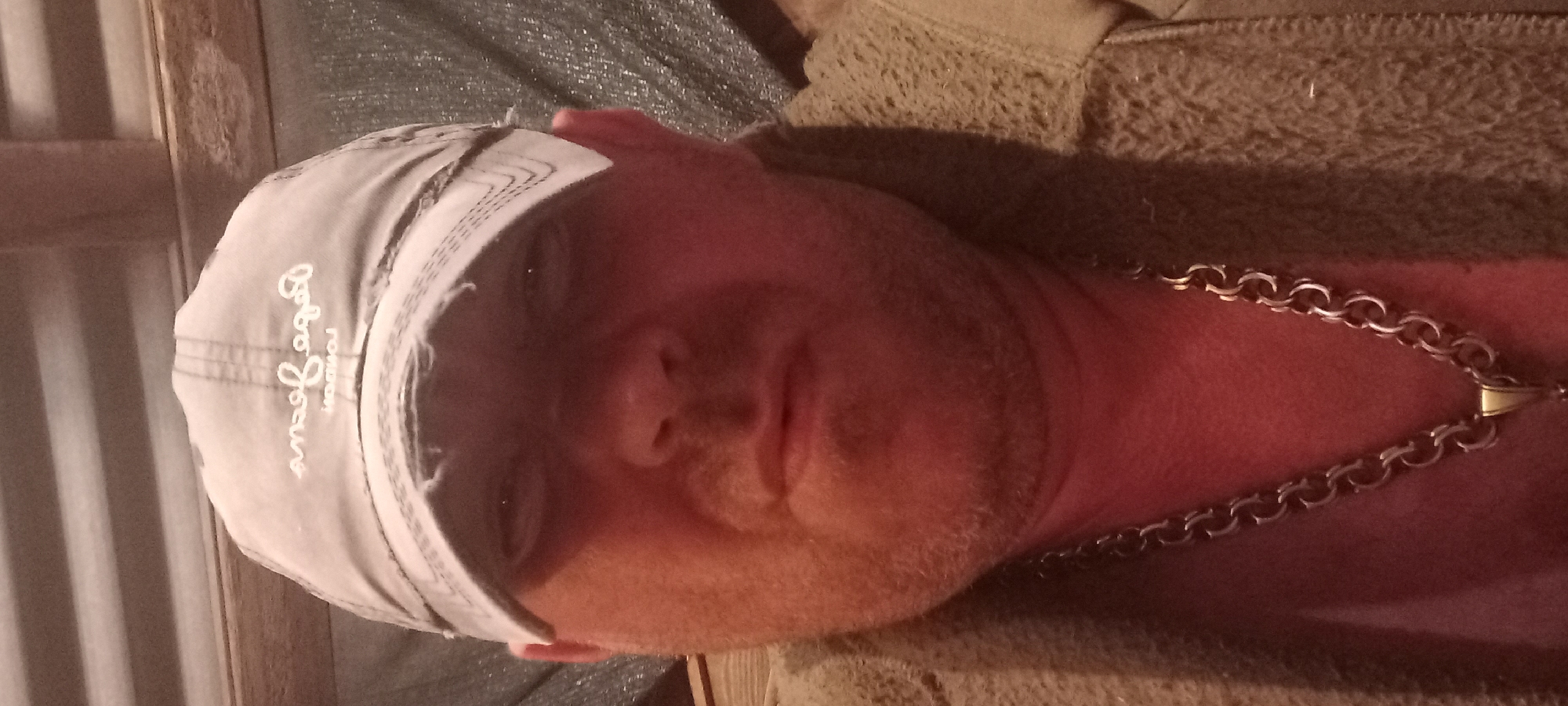 The image presents a close-up, front-facing portrait of a male individual, appearing middle-aged, with fair skin and short reddish-brown facial stubble. He wears a light-colored, possibly grey or khaki, distressed baseball cap with the embroidered text "Pepe Jeans LONDON" visible in reverse on the front. His eyes are partially obscured by the cap's brim, and his expression is neutral. A prominent, thick, silver-colored chain necklace with distinctive round links is visible around his neck, leading to a small gold-colored pendant bail. He is clothed in a heavy-textured, likely brown or khaki-colored garment with a visible collar or lapel of a coarse, woven fabric. The background indicates an indoor, utilitarian setting, featuring corrugated metal sheeting on the upper left and a wooden beam or frame on the upper right. Below the wooden element, a dark, textured material resembling a tarp or fabric is visible. The lighting is warm and somewhat dim, casting a reddish hue on the subject's skin. This scene is located in Kryvyi Rih, Ukraine.