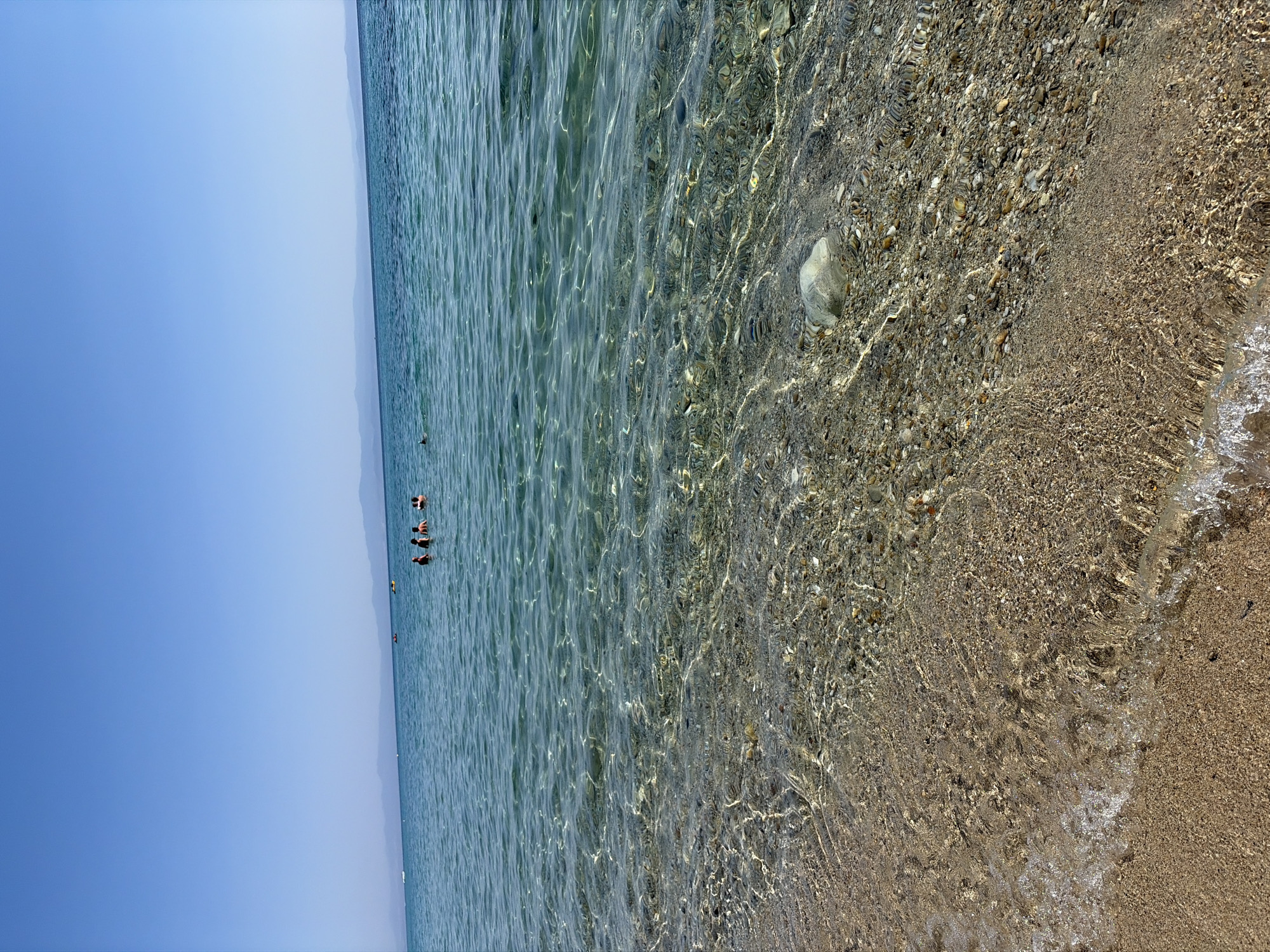 The image captures a shallow section of the sea, likely off the coast of Kassandra Municipal Unit, Greece, during daylight hours. The water is crystal clear, revealing a seabed composed of sand and small pebbles. Sunlight refracts through the water's surface, creating dappled patterns on the submerged ground. In the mid-ground, several people are visible in the water, appearing to swim or stand. The horizon is marked by a distant, hazy coastline with mountains, under a clear blue sky. The foreground shows the sandy beach gently meeting the water's edge, with a small wave lapping at the shore. A large, light-colored stone rests on the seabed near the right side of the frame, partially submerged. The overall scene depicts a serene coastal environment.
