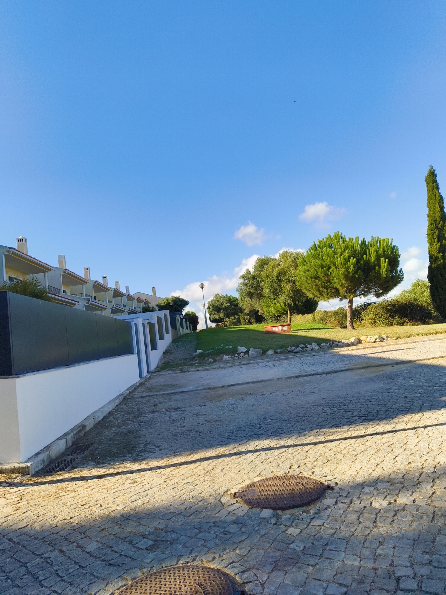The image depicts a residential street in the city of Cascais, Portugal, bathed in the warm glow of afternoon sun. The foreground is dominated by a cobblestone road, with two circular manhole covers visible. A shadow stretches diagonally across the road, cast by an unseen object to the left. To the left of the road, a low, white-plastered wall with a dark grey panel runs along the property line. Beyond the wall, a row of modern, terraced houses with cream-colored facades and red-tiled roofs are visible. Each house features multiple chimneys.
The midground features a grassy slope dotted with rocks, leading up to a line of trees. Prominent among these is a large, rounded pine tree with vibrant green needles, casting a distinct shadow. To its left, other trees with denser foliage are present, and further to the left, a dark green, tall, and slender cypress tree stands prominently. A lamppost with a single light fixture is visible on the grassy slope, between the houses and the trees. The background is a clear, bright blue sky with a few wispy white clouds. A tiny black speck, possibly a bird or an insect, is visible high in the sky. The overall impression is one of a well-maintained residential area on a sunny day.