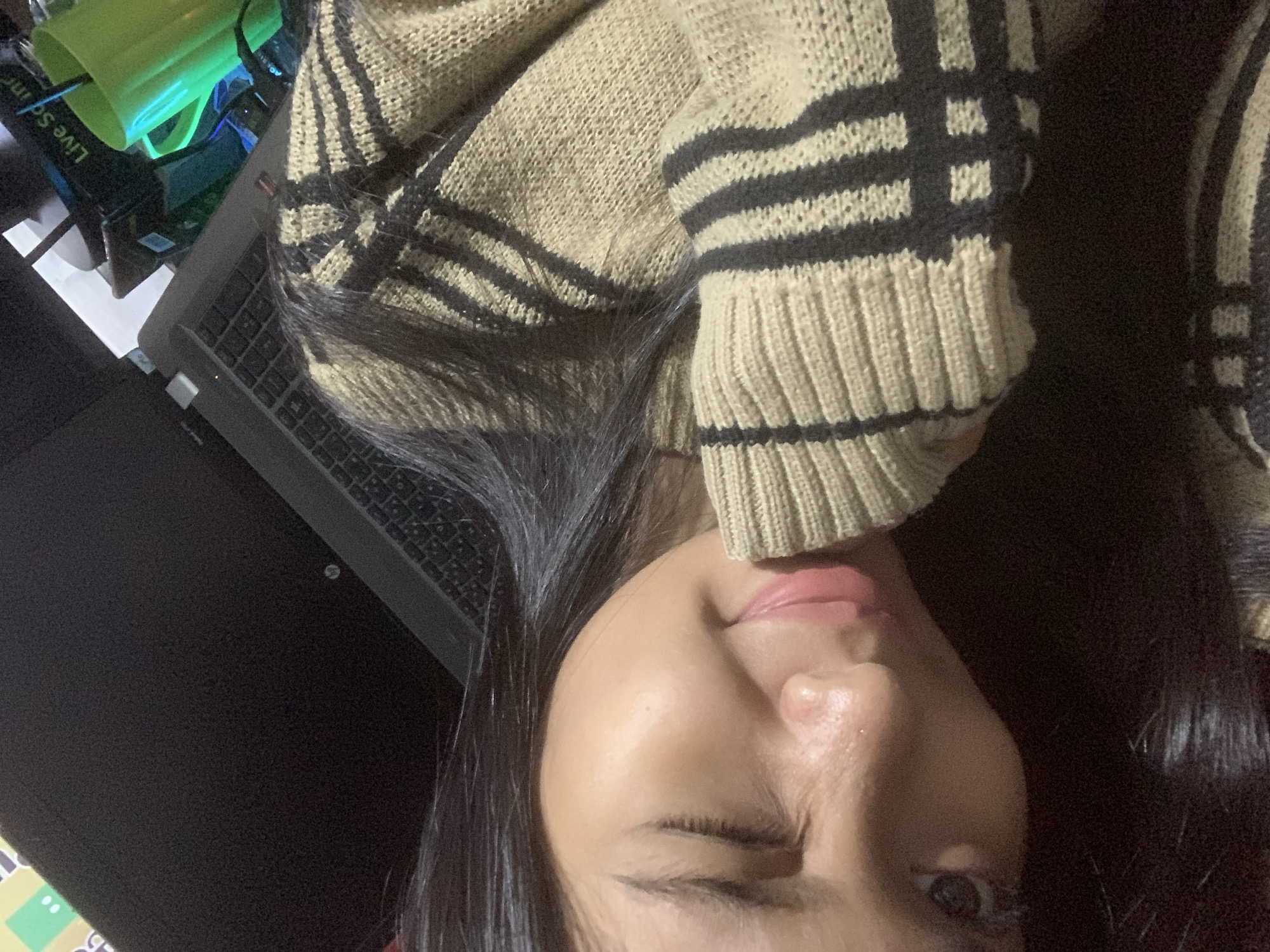 A close-up, slightly skewed photograph shows a person's face, cropped to focus on the right eye, nose, and mouth. The person is wearing a sweater with a tan background and dark stripes. To the left, partially visible, is a laptop computer. In the background are some items including a green container labeled with the word "Live Sound." It is impossible to determine location.