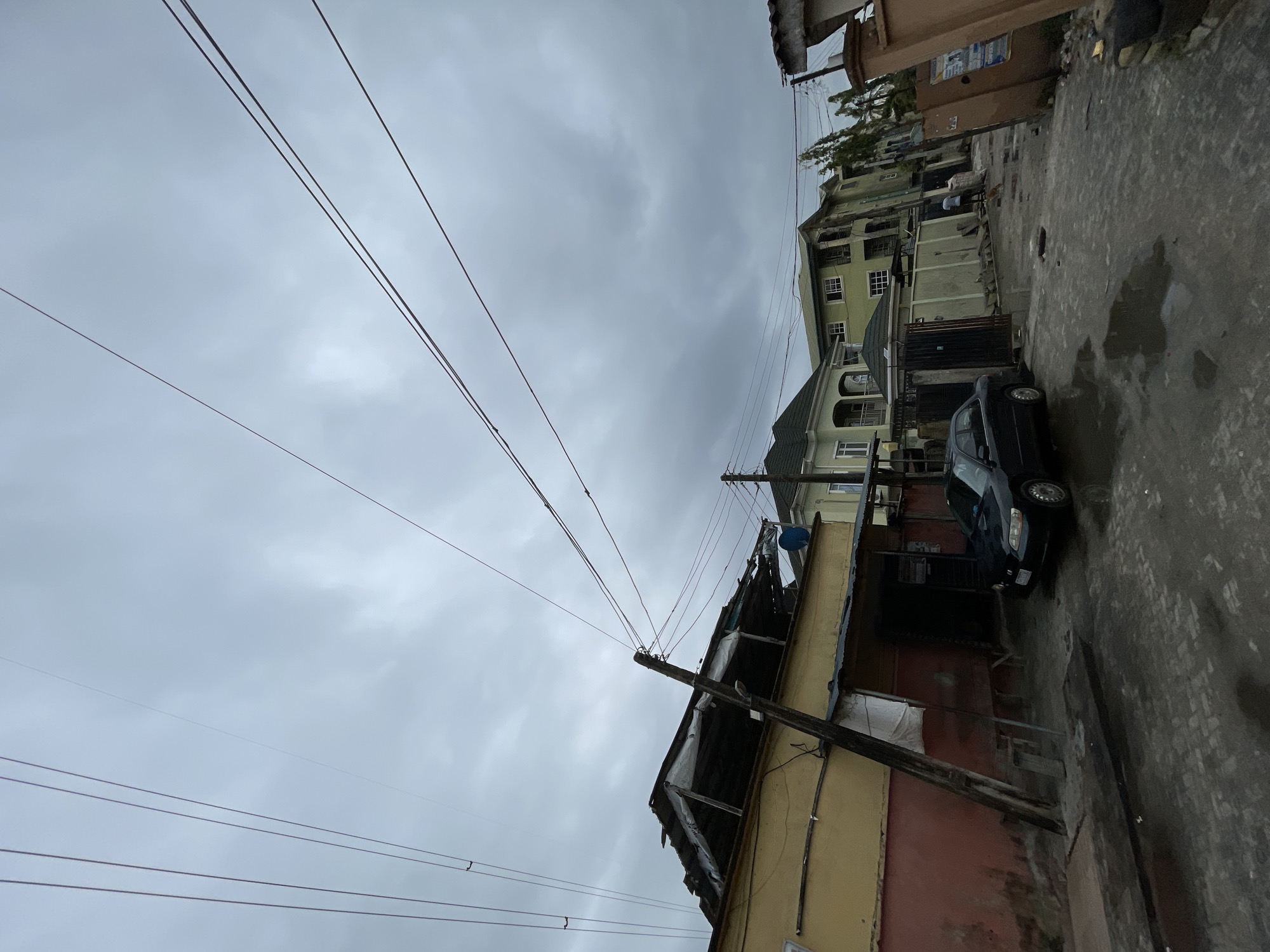 The image captures a street scene in Ado, Nigeria, under an overcast sky. A network of overhead electrical wires crisscrosses the view, supported by wooden poles. A dark-colored car is parked along the side of the narrow street, adjacent to a weathered, single-story building with a dilapidated roof. The building's exterior is painted in a mix of yellow and reddish-brown hues. Further down the street, multi-story buildings with light-colored facades are visible. The pavement is wet, suggesting recent rainfall or damp conditions. There are no people visible in the immediate foreground of the image.