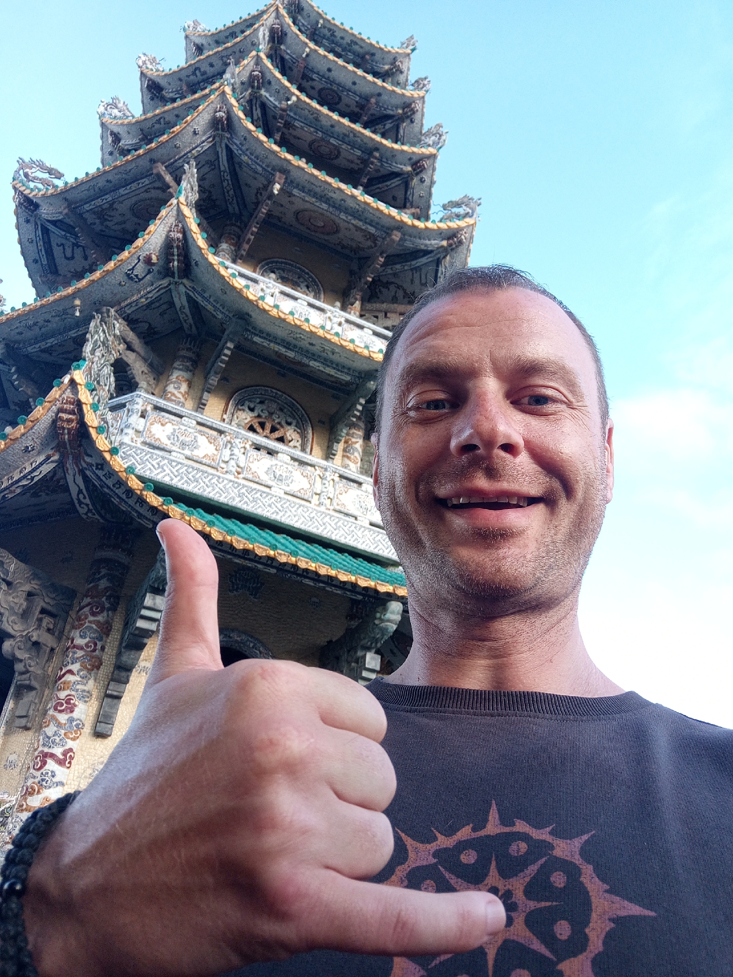 A man with fair skin, short brown hair, and stubble is taking a selfie outdoors, giving a thumbs-up and "shaka" gesture. He is wearing a dark t-shirt with a circular, spiky design. In the background, a multi-tiered pagoda with intricate decorations and green and gold accents is visible against a clear blue sky. The pagoda is located in Xuân Thành, Vietnam, specifically at the Binh An Pagoda, a Buddhist temple known for its unique mosaic-covered architecture. The lighting suggests it is daytime.