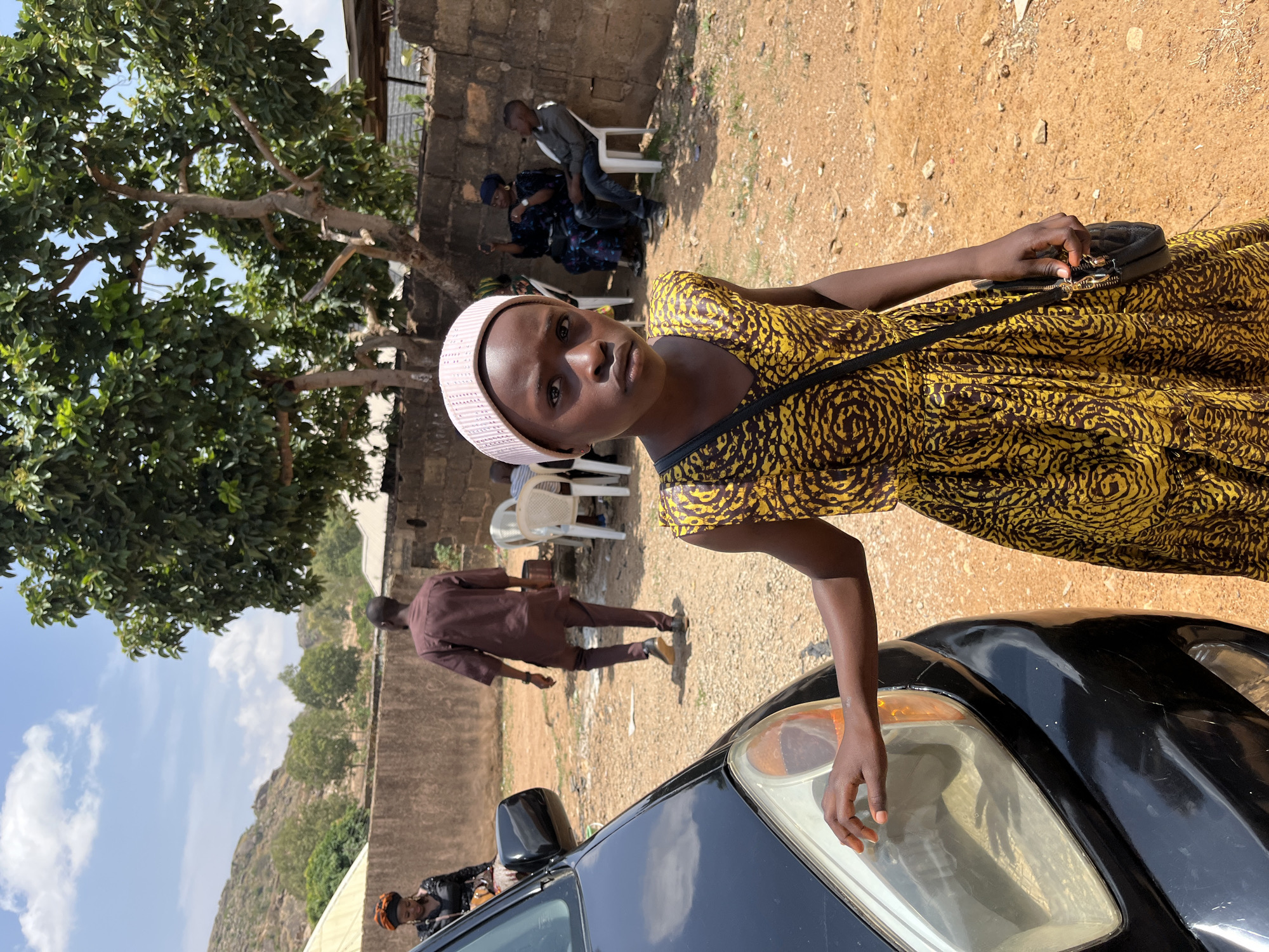 A young woman stands by a black car with her hand on its headlight. She wears a yellow and black dress, a white head covering, and has a black shoulder bag. Behind her, a man in a brown outfit walks away from a group of people seated near a brick wall under a tree. The location is Gura Top, Nigeria, as indicated in the prompt. In the background, a hill is visible under a blue sky with white clouds.