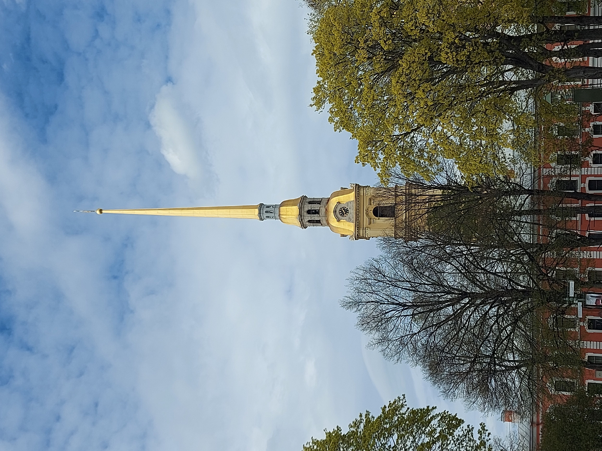 The image shows the Peter and Paul Cathedral in Saint Petersburg, Russia, from a low angle. The gold spire of the cathedral dominates the scene, reaching upwards against a partly cloudy blue sky. The cathedral's building is partially visible behind trees. Some trees have green foliage, while others are bare. A red building with windows is visible behind the trees.