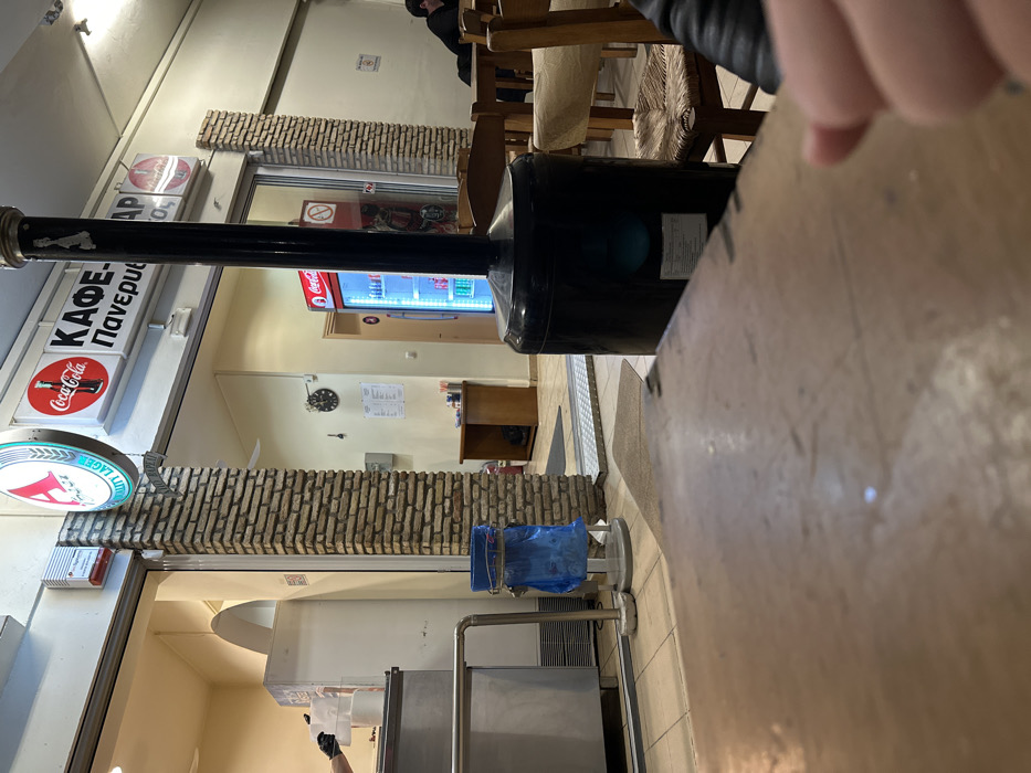 The image depicts the interior of a casual eatery or café, likely in Nea Erithrea, Greece. The perspective is from a table or counter in the foreground, with a person's hand partially visible resting on it.
In the mid-ground, several wooden tables and chairs with woven seats are arranged. A tall, black cylindrical object, possibly a heater or an exhaust pipe, stands prominently. Beyond the seating area, a large, upright beverage cooler displaying the Coca-Cola logo is visible. A smaller wooden cabinet and a wall clock adorn the far wall. Sections of the interior walls are decorated with a brick-like cladding.
Above a doorway, there are multiple signs. A white rectangular sign features the Greek text "ΚΑΦΕ-ΠΑΝΕΜΦ" (KAFE-PANEMF), likely indicating "Café-Panemborio" or a similar name. Below it, a round red sign with the Coca-Cola logo is seen, alongside a green circular sign bearing a white 'A' and the partially visible Greek text "ΛΟΥΤΣ" (LOUTS) and "LAGER", suggesting a beer brand. A "No Smoking" symbol is clearly visible on a glass door.
In the background, near stainless steel commercial equipment, a hand wearing a black glove is present, indicating staff at work. A blue plastic-bag-lined trash bin on a stand is also visible near the counter area. Another person is seated at one of the wooden tables in the mid-background. The overall setting is that of a functional, local Greek establishment, with practical furnishings and clear signage.