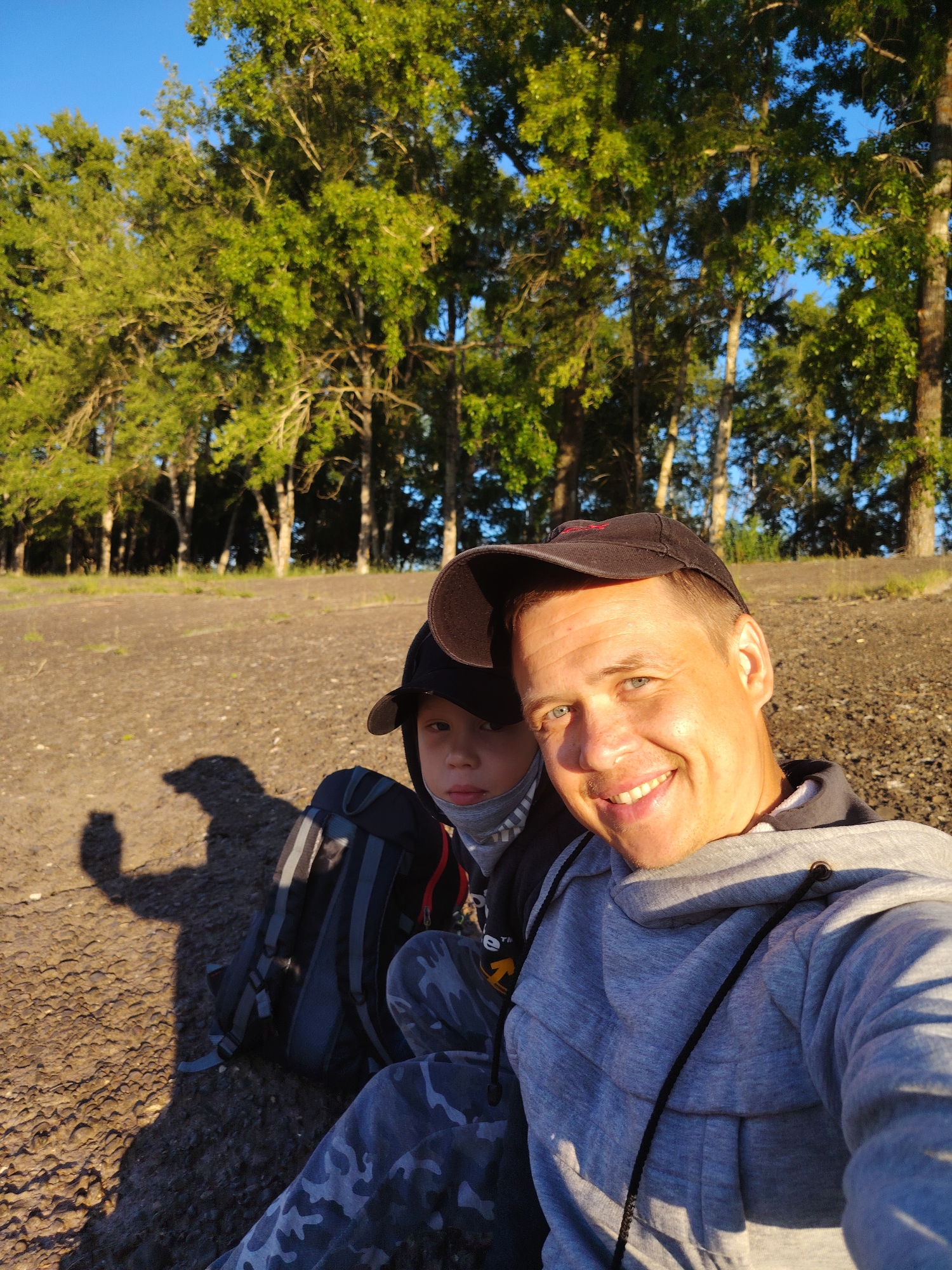 In a sunny, outdoor setting, a man and a boy are taking a selfie. The man, in the foreground, is smiling widely and wearing a gray hooded sweatshirt and a black baseball cap. To his left, partially visible, is a young boy wearing a black hooded jacket, camouflage pants, and a mask. A black backpack with gray straps sits beside them on the gravelly ground. Behind them, a line of tall green trees with lush foliage stretches across the landscape under a clear blue sky. The ground appears to be a mix of dirt and small stones. The scene suggests a recreational outing, possibly in a park or natural area near the Kama River, which flows through Chaikovsky, Russia. The lighting indicates it is likely late afternoon or early morning.