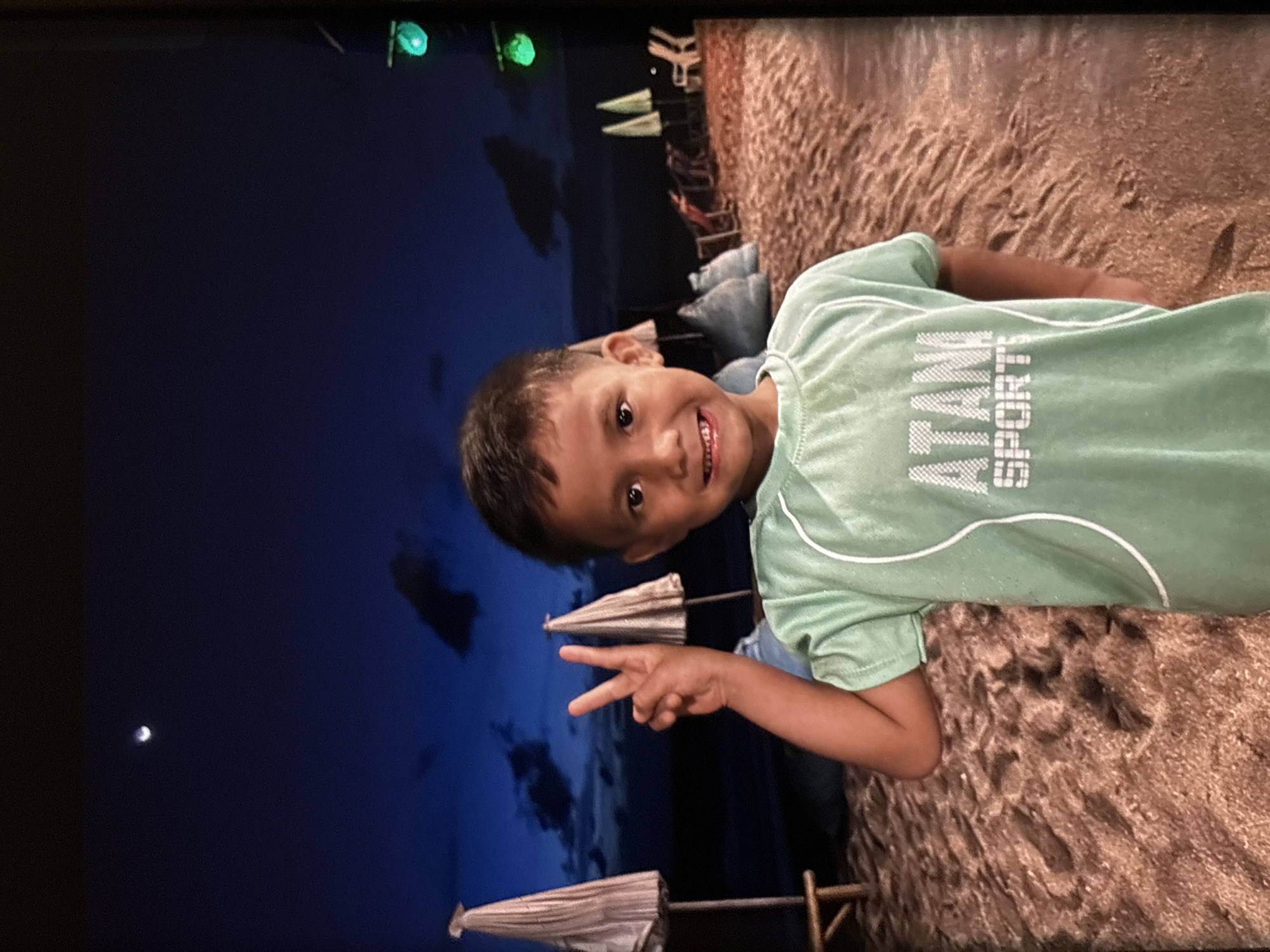 A young boy with dark hair and brown eyes smiles at the camera, holding up two fingers in a peace sign. He is wearing a light green t-shirt with white lettering that reads "ATANI SPORTS". The boy is standing on a sandy beach at dusk. In the background, there are two beach umbrellas, lounge chairs, and several large beanbag chairs. The sky is dark blue with a crescent moon visible. There are lights visible in the distance, some of which are green. The overall scene appears to be a relaxed, tropical evening at a resort or beach club in Chalong, Thailand.