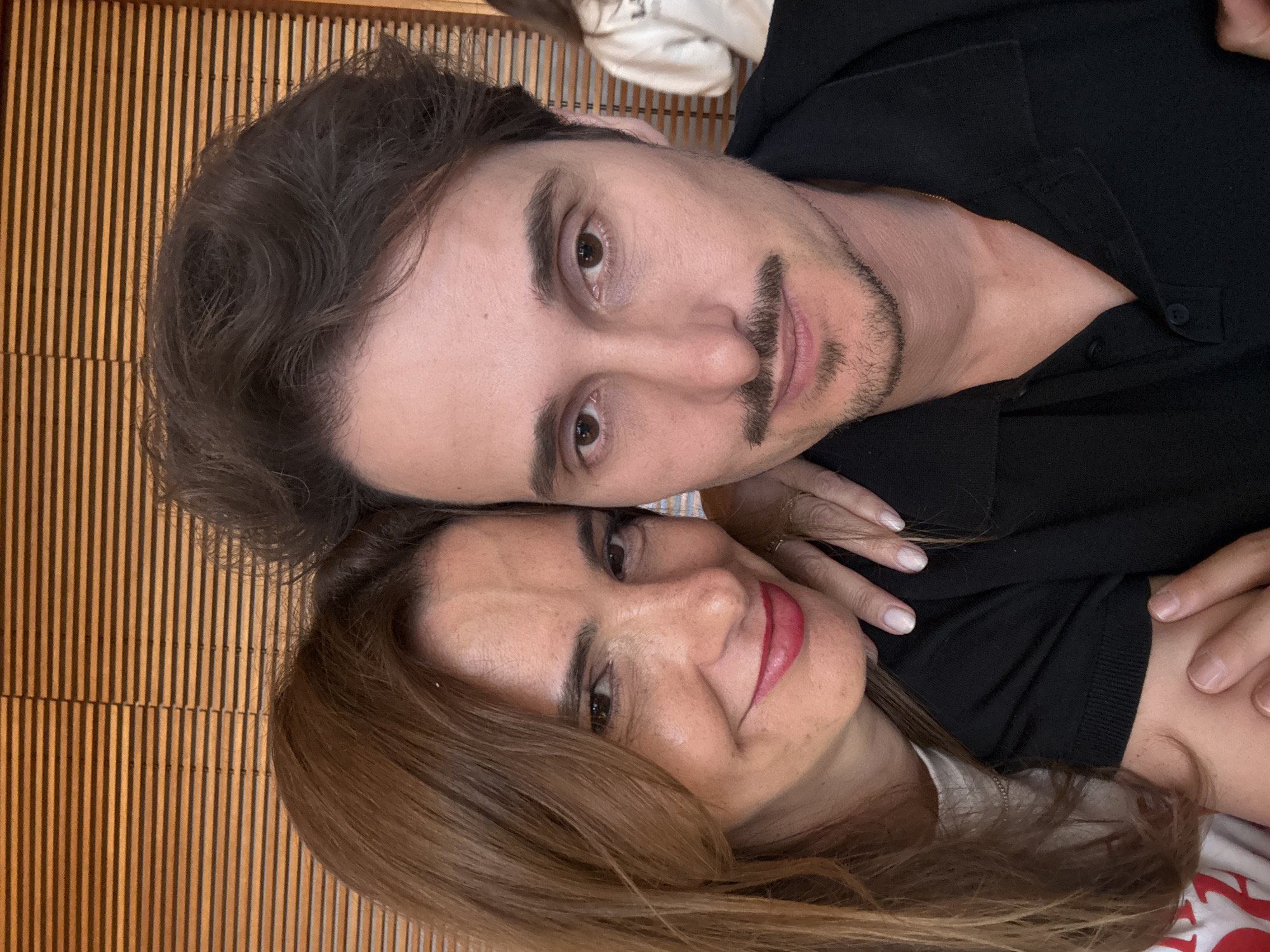 A selfie shows a man and a woman posing closely together. The man has dark brown hair and a dark mustache and stubble. He is wearing a black collared shirt. The woman has light brown hair and is wearing red lipstick. Their faces are close, almost touching. The background appears to be a wall with vertical wood panels. The location of Porto, Portugal is not ascertainable from the image.