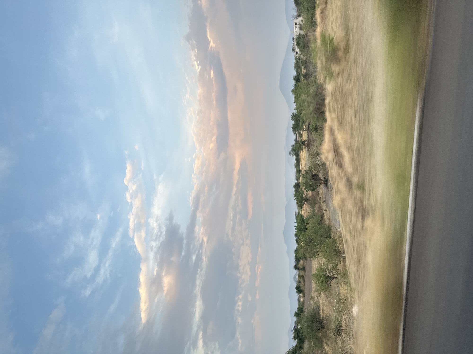The image depicts a landscape view, likely taken from a moving vehicle, possibly in the vicinity of Metepec, Mexico. To the left of the frame, the sky is visible, displaying a mix of light blue and cloud formations. The clouds exhibit a range of shades, including white, gray, and faint orange hues, suggesting possible sunlight reflecting off them.
To the right, the landscape features arid terrain with dry vegetation, trees, and shrubs. A road runs parallel to the viewer, blurring due to the motion of the vehicle. In the distance, faint outlines of mountains can be discerned. The overall environmental conditions appear dry.