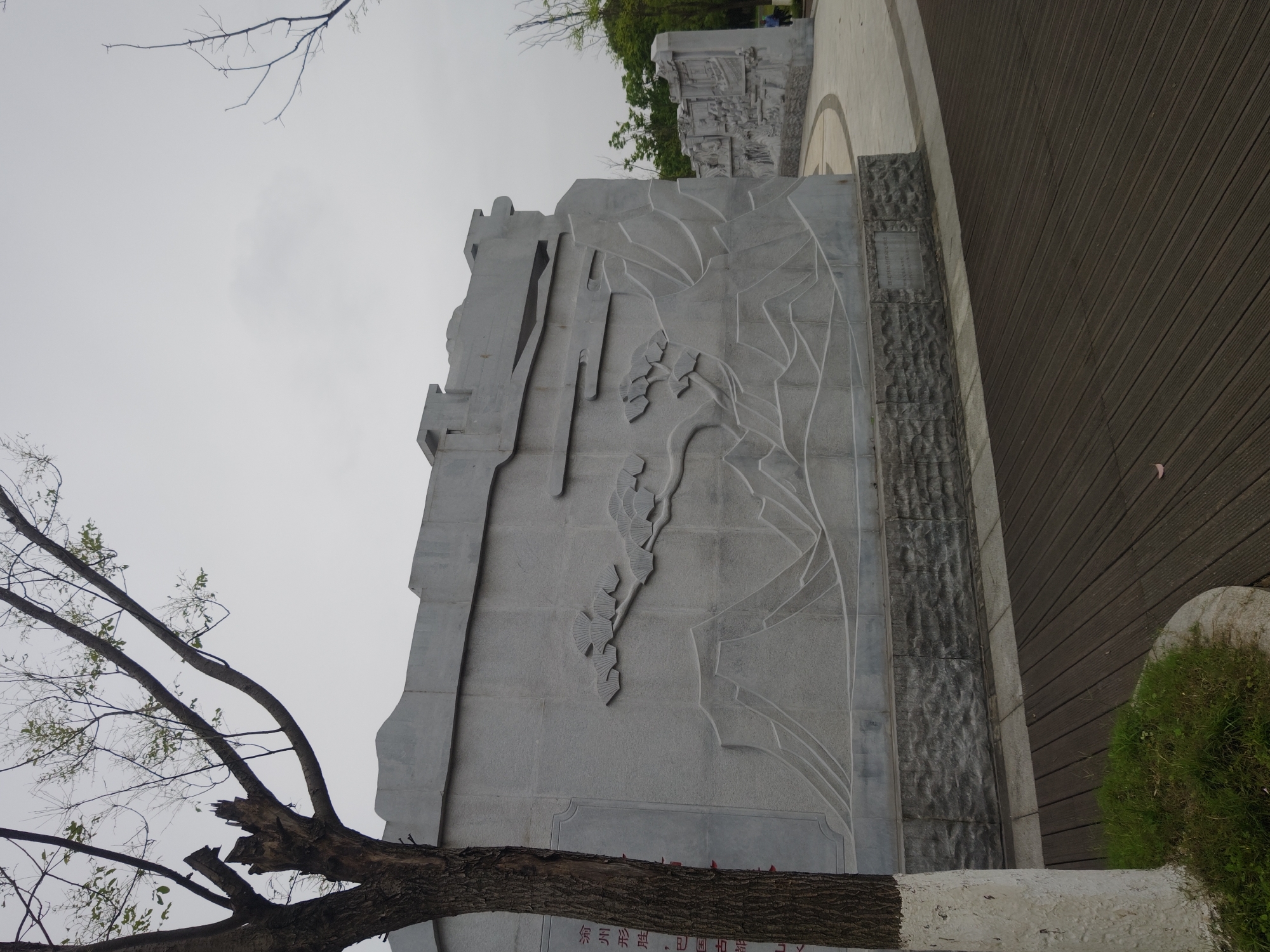 A low-angle, long shot of a gray stone monument or wall in the Chongqing High-tech Zone, China. The wall, which is constructed of large, gray blocks, features a relief carving of a stylized natural landscape, including abstract representations of mountains and a tree branch with fan-shaped leaves. A real tree branch extends across the bottom of the image, obscuring part of the wall. A lighter-colored wooden walkway runs parallel to the wall on the right. In the background, additional stone structures and greenery can be seen. The sky is overcast and gray. The inscription at the base of the monument is partially visible, but the text is illegible.