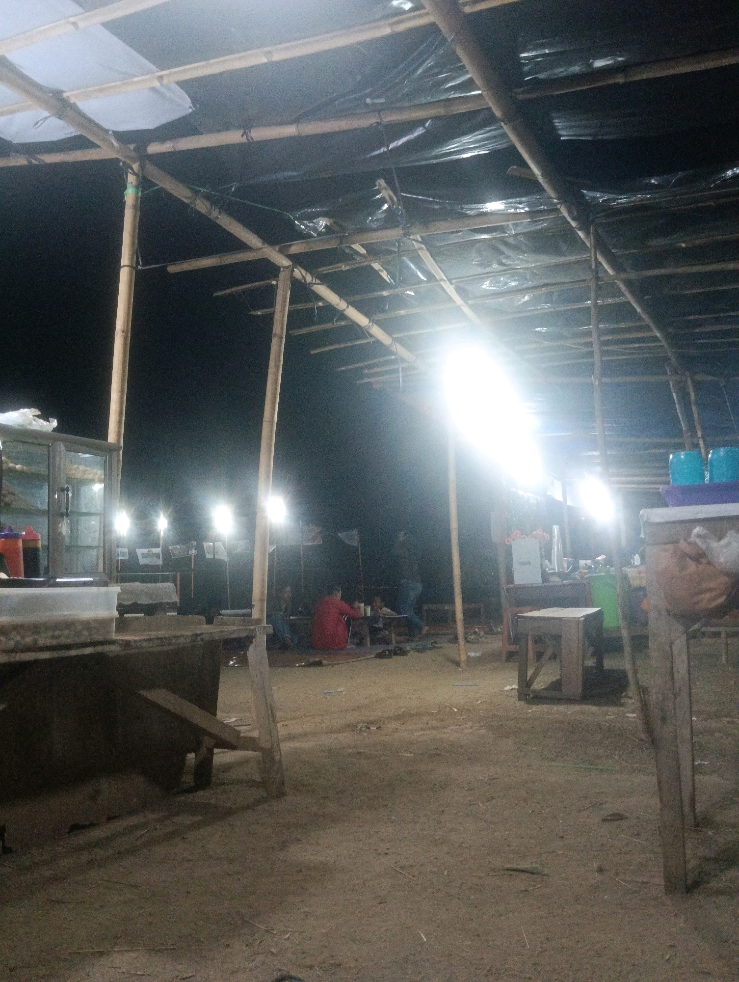This is a nighttime image of an outdoor food stall or marketplace area in Pematang Jaya, Indonesia. The scene is dimly lit by several bright, white artificial lights, some of which are obscured by a bamboo and plastic sheeting roof structure. The ground appears to be dirt or sand.
In the foreground, a substantial wooden counter or table is visible, with its surface blurred and appearing somewhat rough. Behind this, the main area of the food stall unfolds. A glass-fronted display cabinet, likely for food, is positioned to the left, with bottles of sauces on top.
Further back, several people are present, though their features are not clearly discernible due to the distance and lighting. One individual in a red top is seated at a low table. Other figures are either walking or seated in the background, engaged in various activities related to the marketplace. Small stalls or tables are scattered throughout the area.
The overall atmosphere suggests a bustling, informal nighttime gathering, typical of a local market or street food vendors.