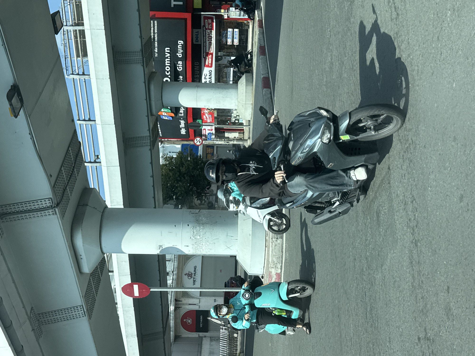 In Thủ Đức, Vietnam, a sunny day shows several people riding motorbikes on a paved road under an elevated structure. A person wearing a turquoise jacket and a face mask rides a light blue electric scooter in the foreground, on the left side of the frame. Behind them, another person on a white motorbike is partially visible. Further back, a person on a dark grey motorbike rides in the center of the image, wearing a black hoodie and a face mask. The elevated structure features concrete pillars and metal beams, with some advertisements visible on a building in the background. The road shows tire marks and reflections from the sunlight.