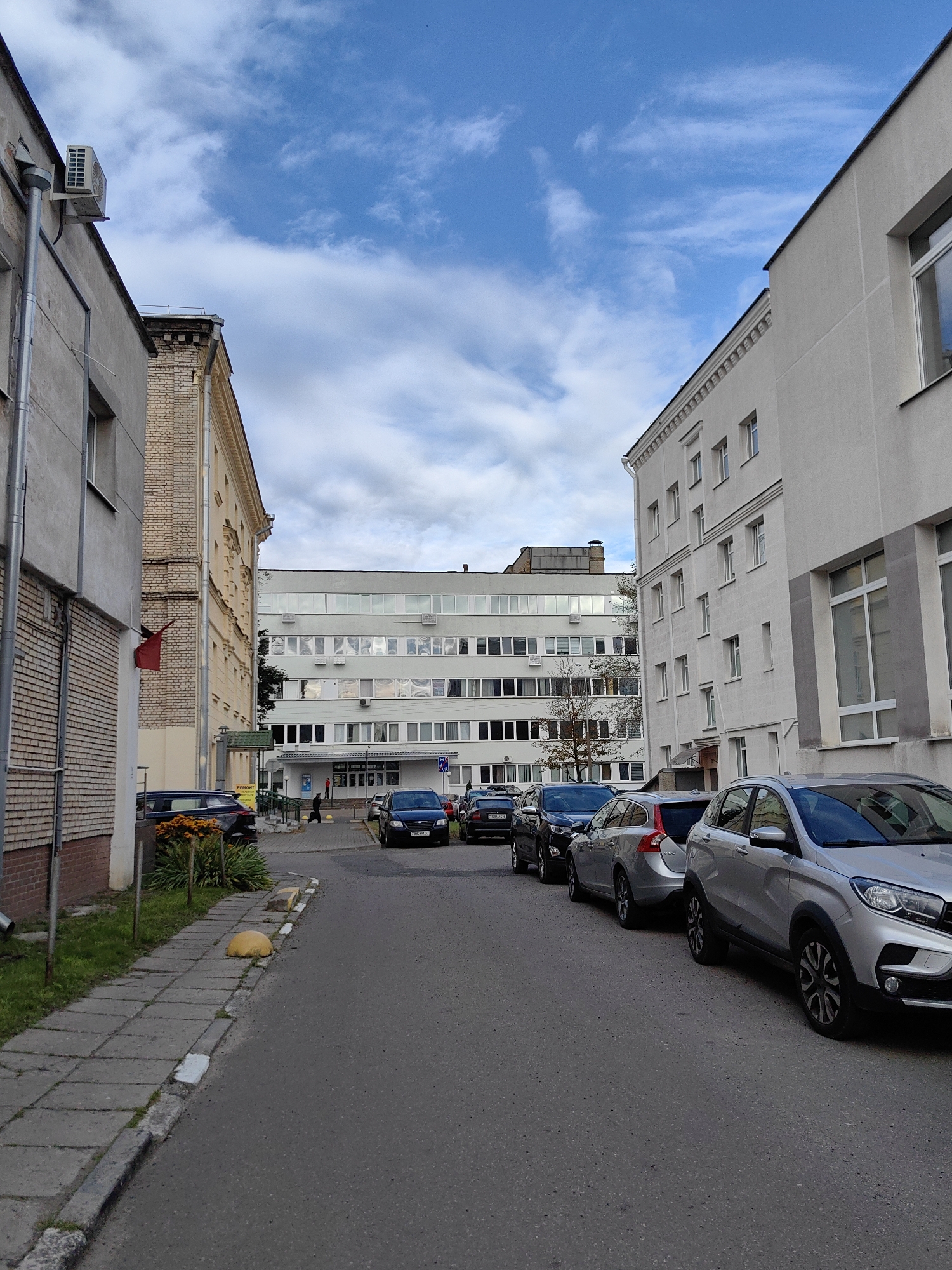 In an urban scene, likely in Minsk, Belarus, a street lined with parked cars runs between two buildings. On the left, a light-colored brick building with a grey facade and a prominent air conditioning unit is visible. Further down, a lighter yellow building with ornate architectural details stands adjacent. A red flag is affixed to a pole near the yellow building.
On the right, a modern, multi-story white building with numerous windows dominates the view. The street itself is paved with asphalt and features a grey concrete sidewalk on the left, bordered by grass and a yellow parking bollard. Several cars are parked along the right side of the street, including a silver SUV in the foreground. In the middle ground, a larger, multi-story building with rows of windows is visible, and a few pedestrians are walking near its entrance. The sky above is blue with scattered white clouds.